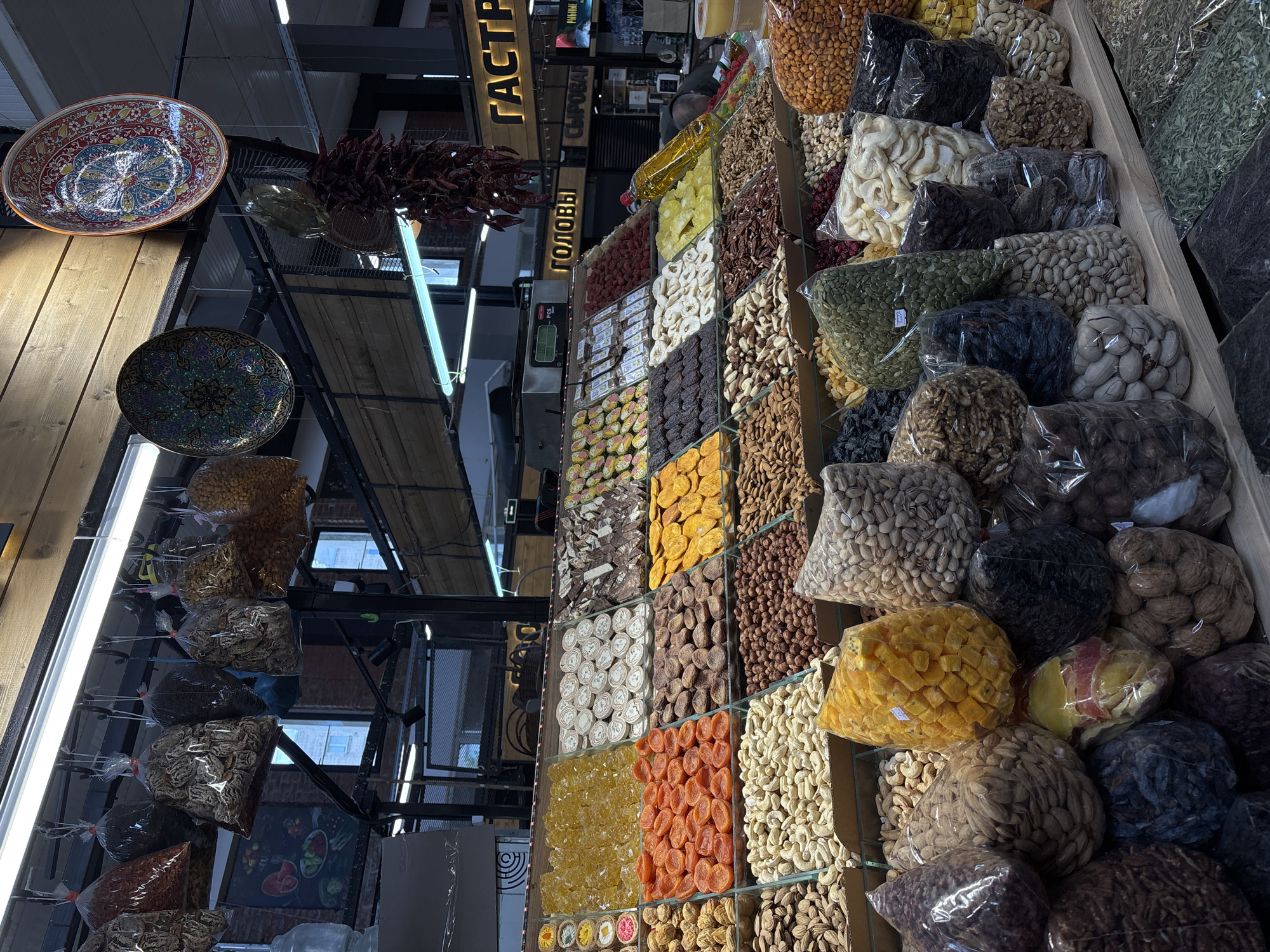 The image shows a market stall with a variety of dried fruits and nuts for sale. The stall is located in Saint Petersburg, Russia, based on the Cyrillic signage visible. The dried fruits and nuts are displayed in clear plastic bags arranged in rows. Varieties include apricots, raisins, walnuts, cashews, and pistachios. Above the stall are hanging decorative plates and dried chili peppers. Overhead, the market structure is visible, including metal beams and lighting fixtures. The stall is well-lit.