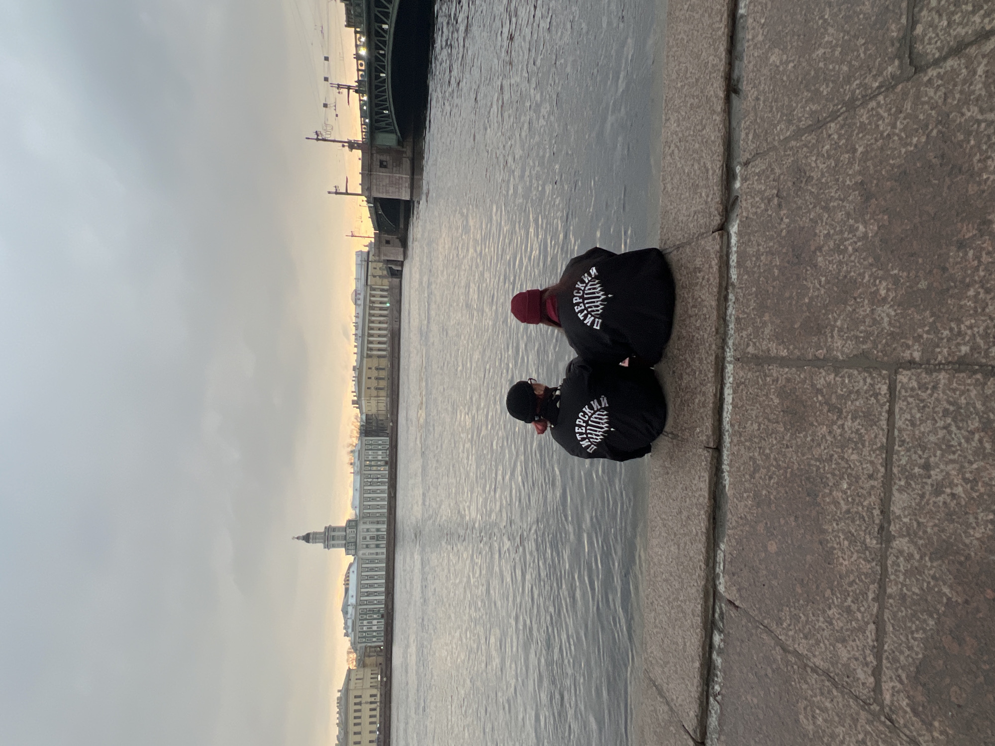 Two individuals are seen sitting on a stone embankment, with their backs to the viewer. Both are wearing black jackets with white text on the back. One person has a red beanie. The background shows a body of water, likely a river, reflecting the sky. On the far side of the water is a cityscape, including a tall, thin tower and a bridge structure. The image is taken in Saint Petersburg, Russia, based on the architectural style and river embankment.