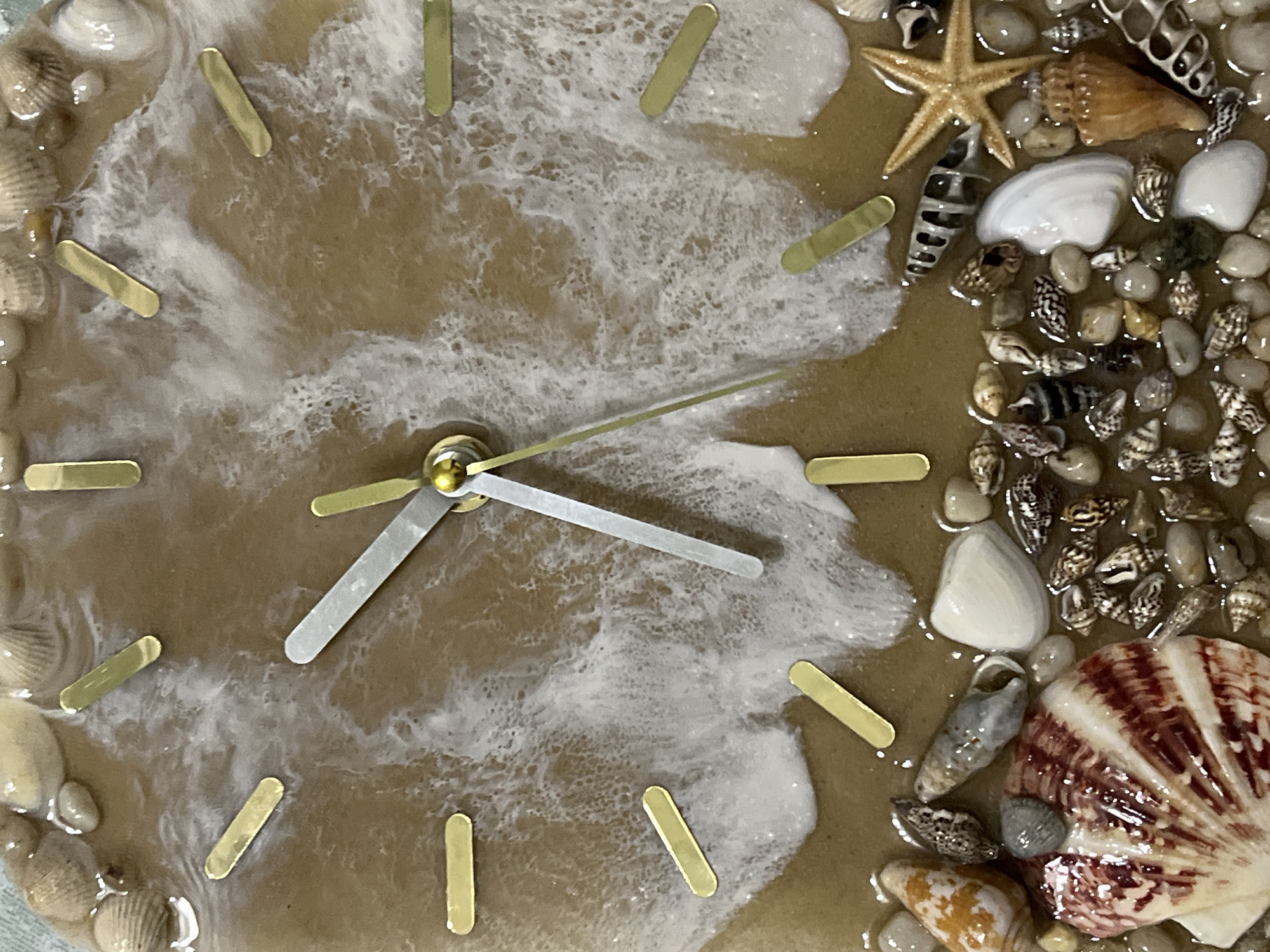 The image is a close-up, overhead shot of a decorative clock face. The clock is designed to resemble a beach scene, with a sandy brown background and white resin "waves." There are gold rectangular hour markers around the perimeter of the clock face. The clock has silver hour and minute hands, with the hour hand pointing approximately to the 11 and the minute hand pointing to the 4. The bottom portion of the clock is adorned with a collection of seashells, pebbles, and a small starfish. The specific location of this clock in Belyye Berega, Russia, cannot be determined from the image.