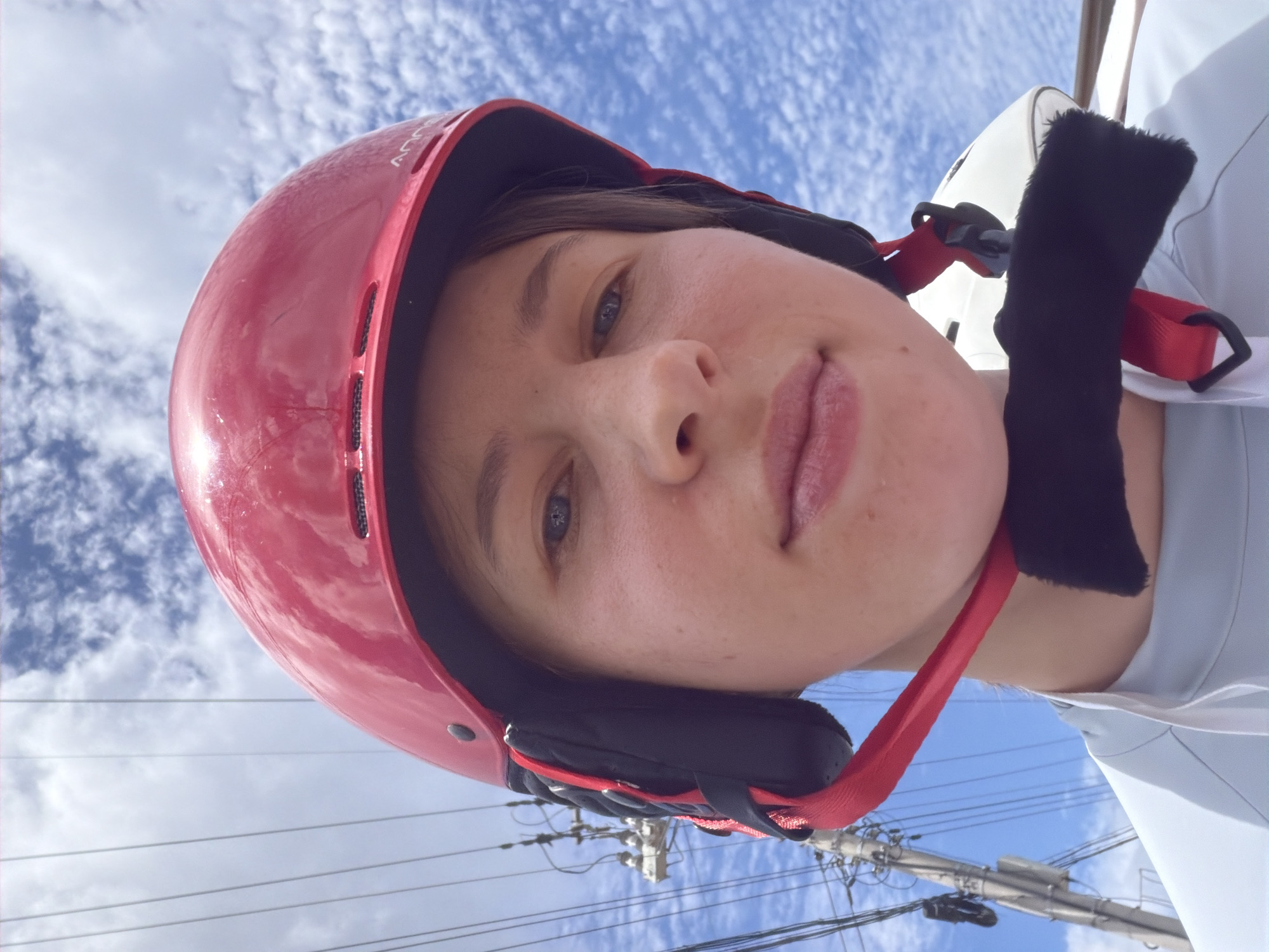 A close-up, selfie-style photograph shows a person from the chest up, wearing a red helmet with black ear pads and a red chin strap. The person has fair skin, light-colored eyes, and short brown hair visible around the helmet. The background is a bright blue sky with wispy white clouds. To the left, a utility pole with numerous wires is visible. The person appears to be outdoors. The image does not contain any specific geographical markers that would indicate it was taken in Seo-myeon, South Korea.
