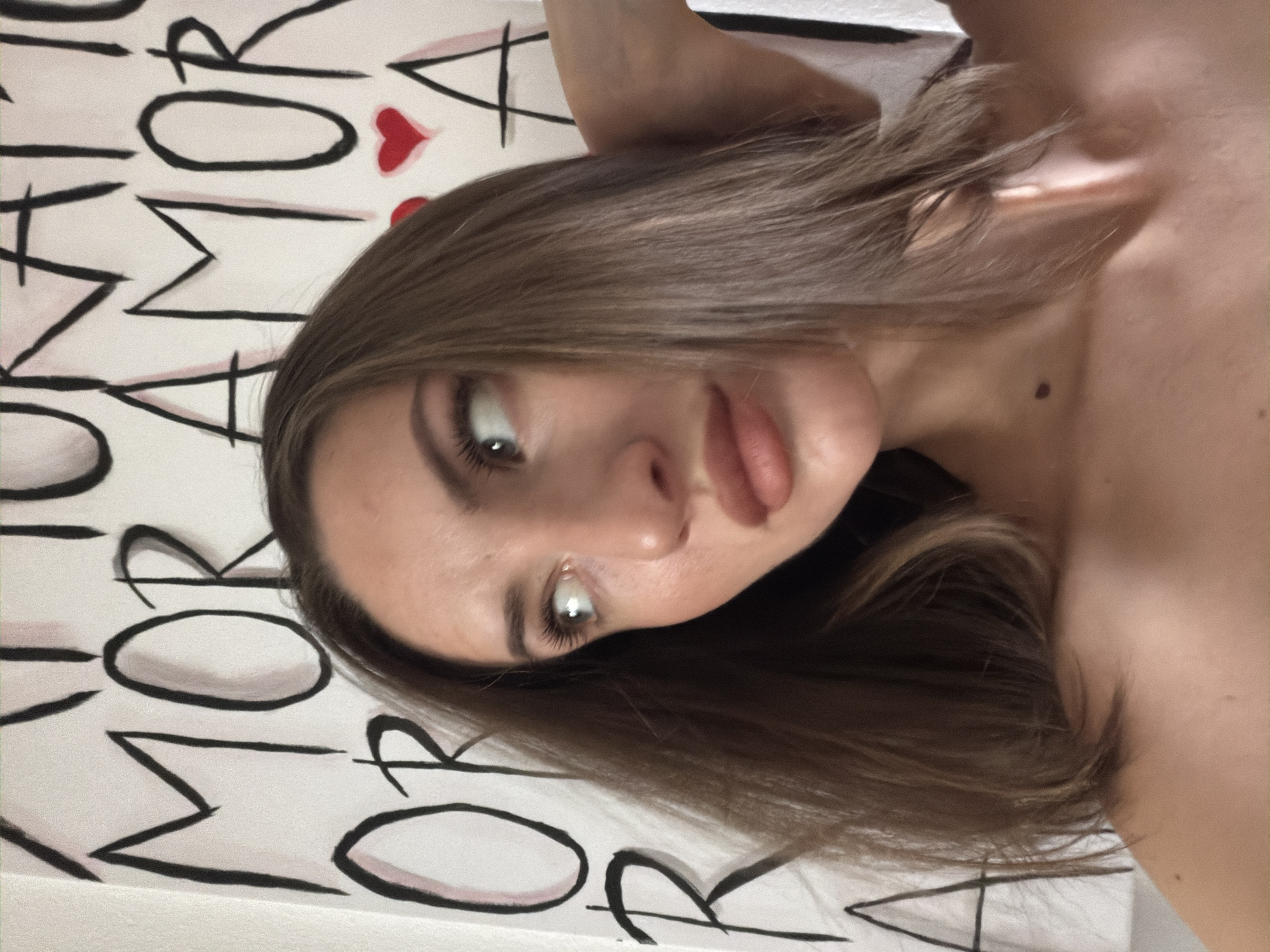 The image is a medium shot of a young woman with brown hair, looking to the right. She is in front of a white wall with large black lettering and red hearts painted on it. The text appears to be in Italian and says "AMOR AMOR" and other words. The woman is wearing makeup, including mascara and lipstick. Her left arm is raised and resting on the back of her head. The lighting is natural and soft. The background suggests an indoor setting, possibly a studio or a room with a painted mural. There is no information in the image to indicate the location is Sharm El Sheikh, Egypt.