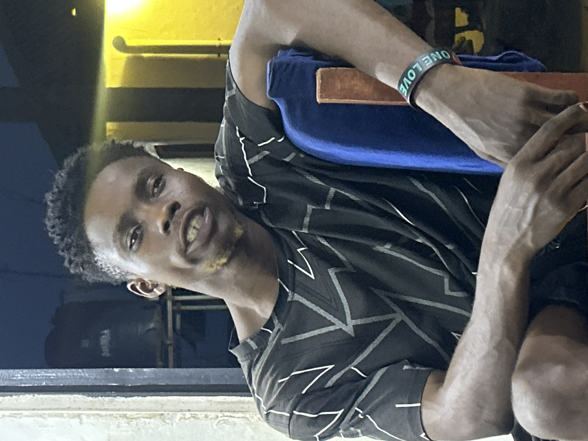A medium shot, taken outdoors during the day in Unknown, Unknown, features a dark-skinned man wearing a dark gray patterned shirt. He has short, dark hair and facial stubble. He is positioned behind a light-colored ledge, possibly a windowsill. Behind him and to the left, there is a dark blue surface, possibly a wall or tank. Above him, there is a yellow-toned structure, possibly part of a building. He is seated in a chair and has a blue pillow that is supported by a brown-colored frame. The man is wearing a dark-colored wristband with light-colored writing on his right arm that reads "ONE LOVE".