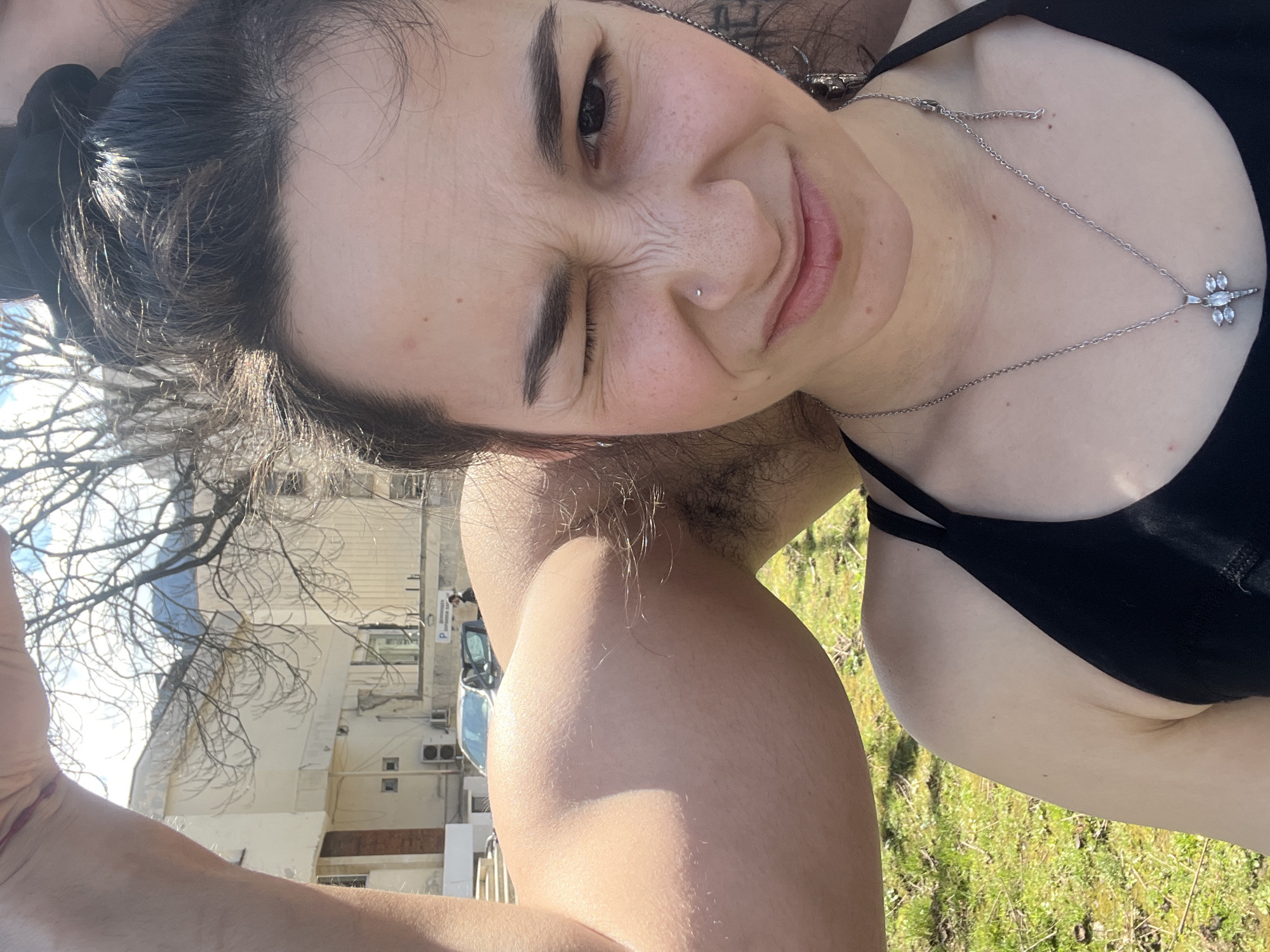 A woman with dark hair pulled back in a bun is taking a selfie outdoors. She is winking and smiling slightly. She has a nose piercing. She is wearing a black top and a silver necklace with a dragonfly pendant. Her arm is raised in the foreground. The background shows a building with bare trees, a parking space, and a car. The location is possibly Vratsa, Bulgaria. Grass is visible on the ground.