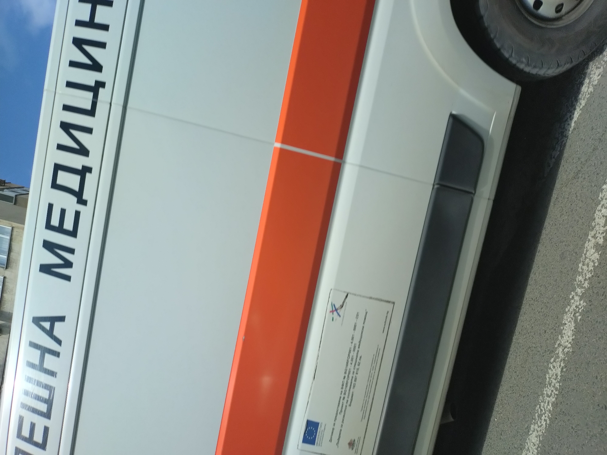 An upward-angled, tilted shot captures the side of a white van, likely an ambulance, under a clear blue sky during daylight. The van's side is dominated by a prominent orange vertical stripe. Bold black Cyrillic text, reading "СПЕШНА МЕДИЦИН" (part of "СПЕШНА МЕДИЦИНА," meaning "Emergency Medicine"), is visible on the front section of the van.
Below the orange stripe, a rectangular information plaque is affixed. It displays the European Union flag and another official emblem, suggesting a funded project. The text on the plaque, in Bulgarian, details:
- "Проектът е финансиран по ОП „Регионално развитие“ 2007-2013" (The project is financed under OP "Regional Development" 2007-2013).
- "Договор за предоставяне на БФП № BG161PO001-4.2.01-0002" (Grant agreement for financial aid No. BG161PO001-4.2.01-0002).
- "Спешен център – Спешна медицинска помощ" (Emergency Center – Emergency Medical Aid).
- "Обща стойност на проекта: 1 500 000 лв." (Total project value: 1,500,000 BGN).
- "бюджет: 150 487, 015, 62 лв." (Budget: 150,487,015.62 BGN).
Part of a black side trim or bumper is visible below the plaque. In the foreground, the van's wheel with a dark tire is seen, along with a portion of an asphalt surface marked with a faded white line. In the distant background, a building is partially visible. The scene depicts a stationary emergency vehicle, likely in Stara Zagora, Bulgaria, given the linguistic and contextual cues.