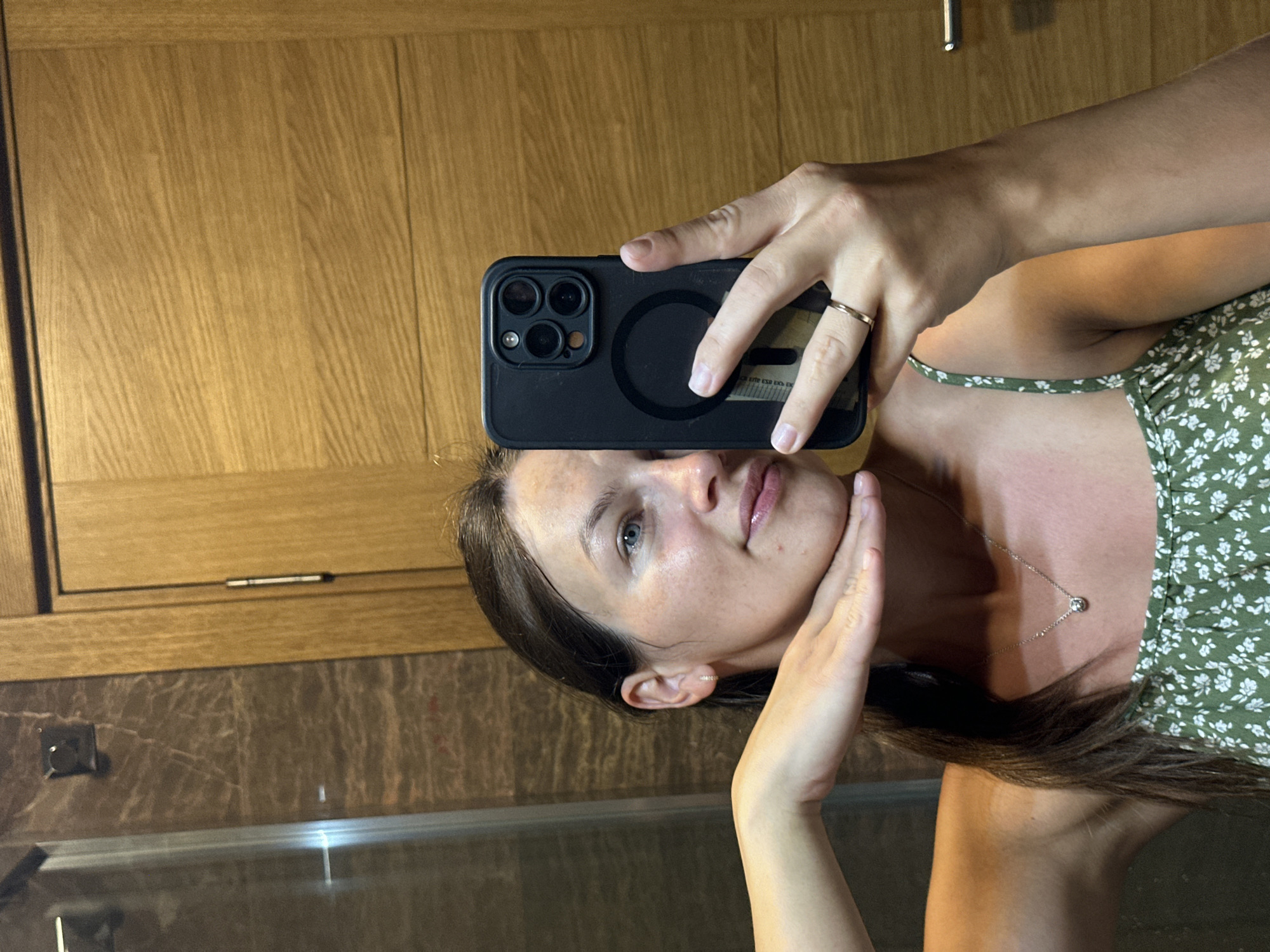 A young woman is taking a mirror selfie in what appears to be a bathroom. She has brown hair pulled back in a ponytail and is wearing a green floral print dress. Her left hand is raised to her chin, and she is holding a black smartphone in her right hand, which is also wearing a gold ring. The background shows a tiled wall with a marble pattern and a wooden door. The overall lighting is warm and natural. The scene is likely in a residential setting.