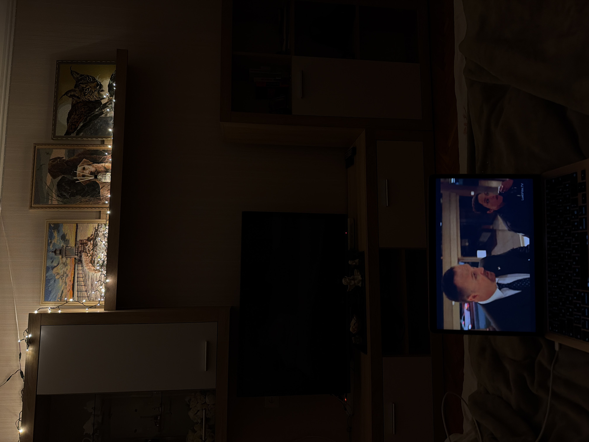 The image depicts an indoor scene, likely a living room, at night. A laptop is open in the foreground, displaying a television show with two actors visible on the screen. The room is dimly lit, with string lights illuminating a shelf above a television. The shelf displays three framed pictures: one of a lighthouse, another of dogs, and a third of a lynx. To the left of the television, a large cabinet with glass doors is partially visible. To the right, a tall cabinet with shelves is also visible. The overall ambiance suggests a relaxed evening of entertainment. The location is identified as Cheltuitori, Moldova.