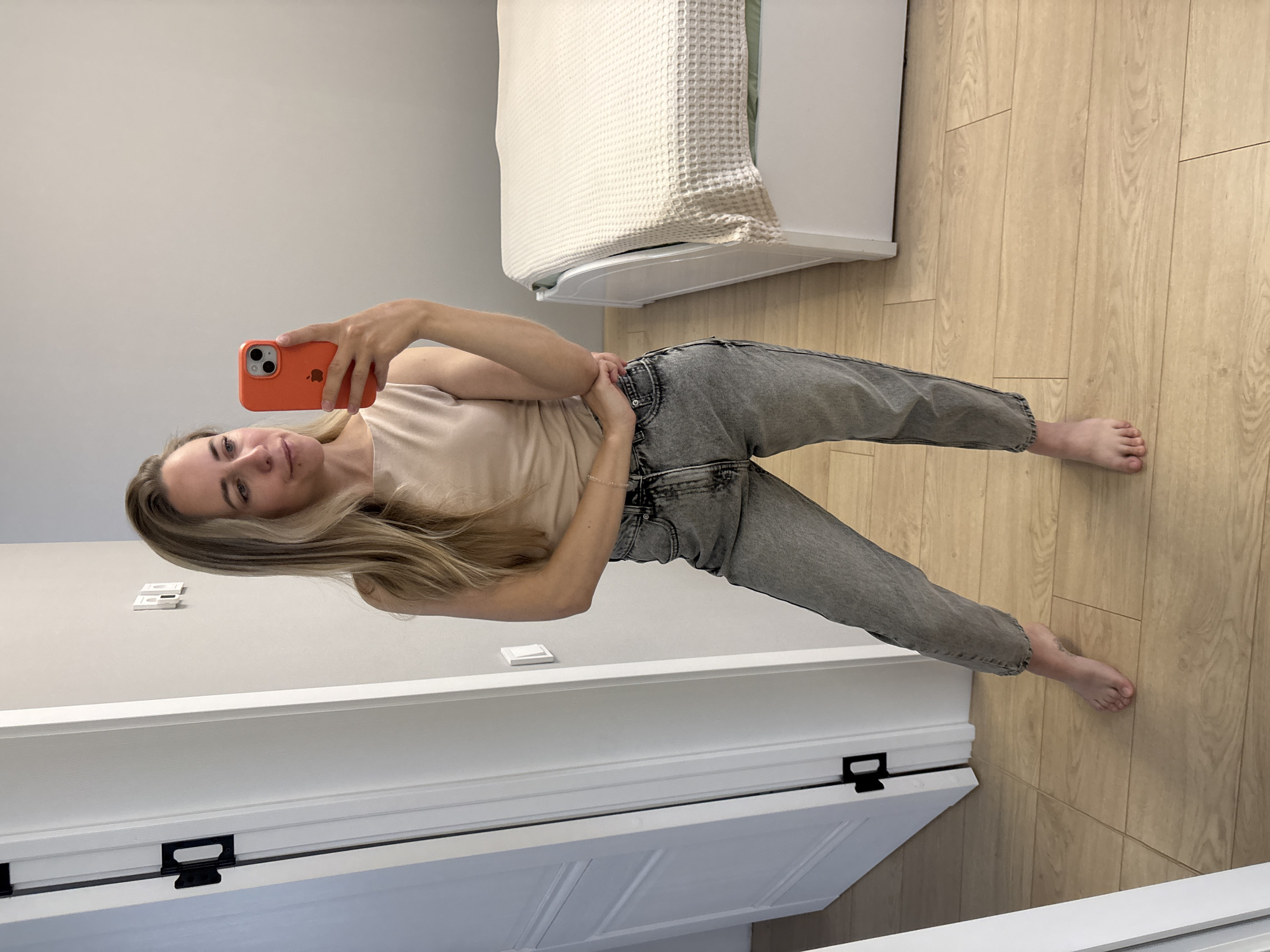 A woman with blonde hair is standing in a room, taking a selfie with her phone. She is wearing a light-colored tank top and grey jeans. Her left arm is bent and resting on her hip, while her right hand holds her orange phone. The room has light-colored walls and wooden floors. To the left, there is a white door with black hardware. To the right, a bed with a white textured comforter is visible. The image appears to be taken in a bedroom or living space.