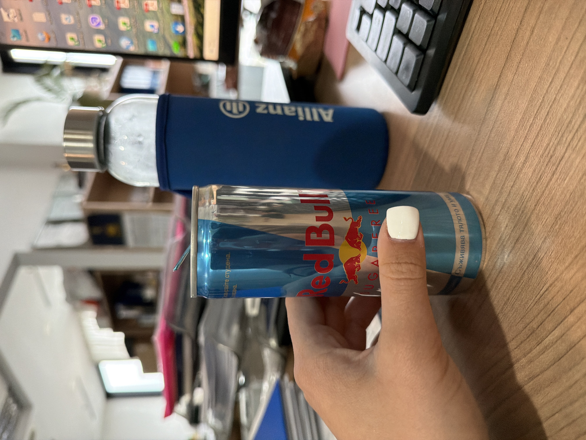 A partially sideways, close-up shot depicts a hand holding a Red Bull sugar-free can against a background consisting of an "Allianz" water bottle, a computer keyboard, a computer monitor, and what appears to be office furniture. The can is mostly silver with blue accents and the distinctive Red Bull logo. The hand has a white nail polish on at least one fingernail. The Allianz bottle is covered in a blue sleeve. The setting is possibly an office in Sofia, Bulgaria, given the text on the can being in Bulgarian.