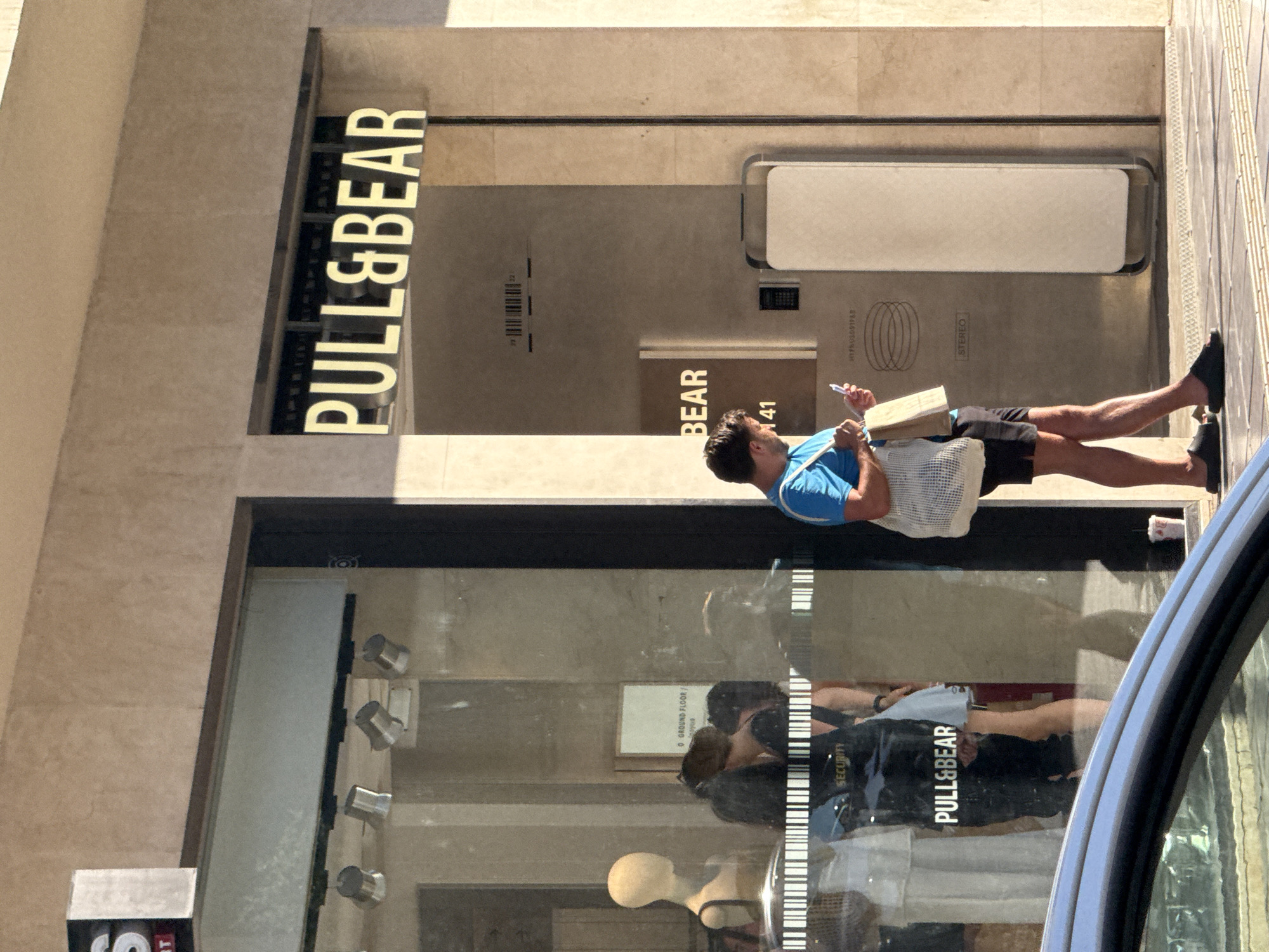 A man in a blue t-shirt and black shorts stands on a sidewalk in front of a store in Chania, Greece. He is holding a phone in his right hand and a paper bag in his left. The store, "PULL&BEAR," has a large glass window displaying mannequins. A silver car is parked in the foreground. The scene appears to be during the daytime, with sunlight casting shadows.