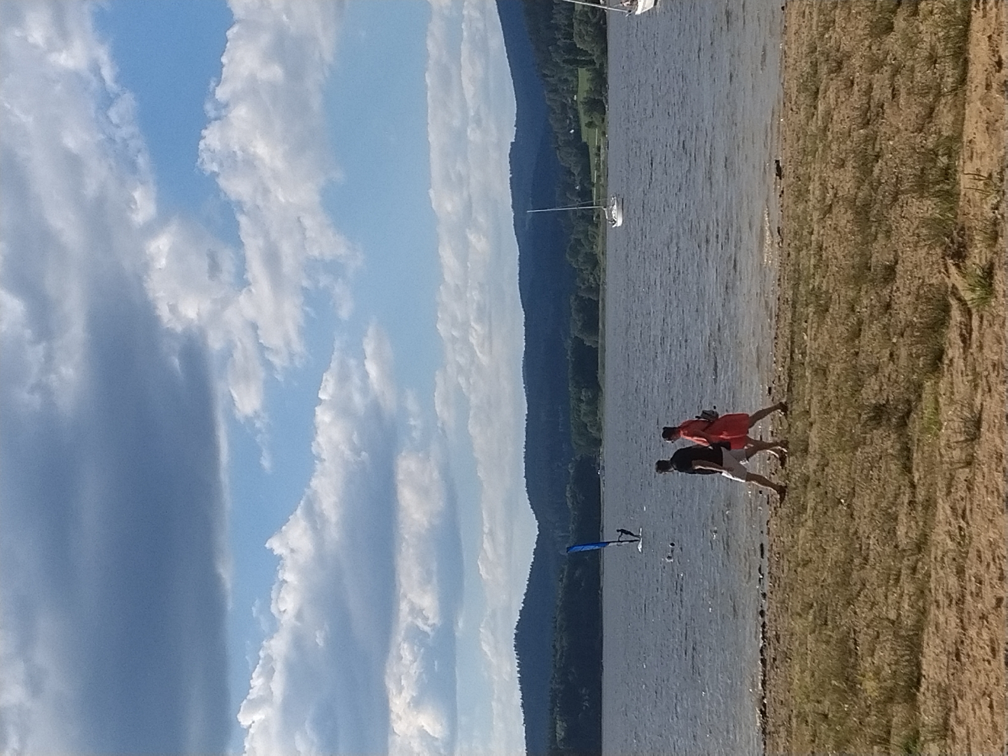 In Radslav, Czechia, on a body of water with dark blue, choppy waves, a person is windsurfing with a bright blue sail. In the distance, a white sailboat with a tall mast is visible on the water. The shoreline features a sandy beach with scattered dry grass and small plants. Two people are walking along the shoreline; the person on the left wears a black t-shirt and white shorts, while the person on the right is in a red dress. The background consists of lush green, tree-covered hills under a sky with dramatic, dark gray and white clouds interspersed with patches of bright blue.