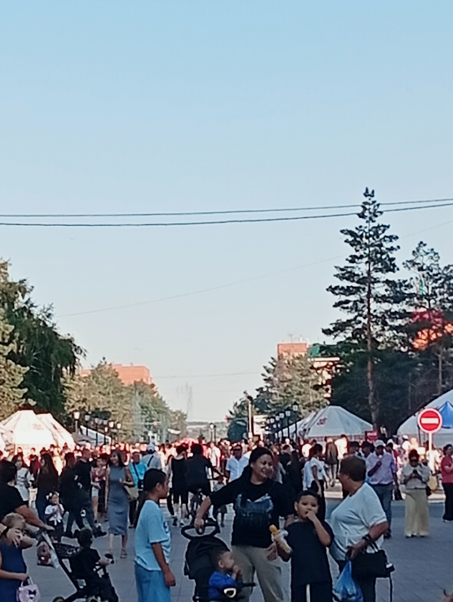 The image depicts a crowded street scene, likely a public thoroughfare or promenade, in Kostanay, Kazakhstan. The sky is clear and pale blue. Several tall pine trees are visible, interspersed with deciduous trees and buildings. Numerous people are walking, some pushing strollers. A red and white circular traffic sign, indicating "no entry" or a similar prohibition, is prominently displayed on the right side of the image. Tents or market stalls are set up along the sides of the street, suggesting a fair or event. The overall atmosphere appears to be one of public gathering and activity.