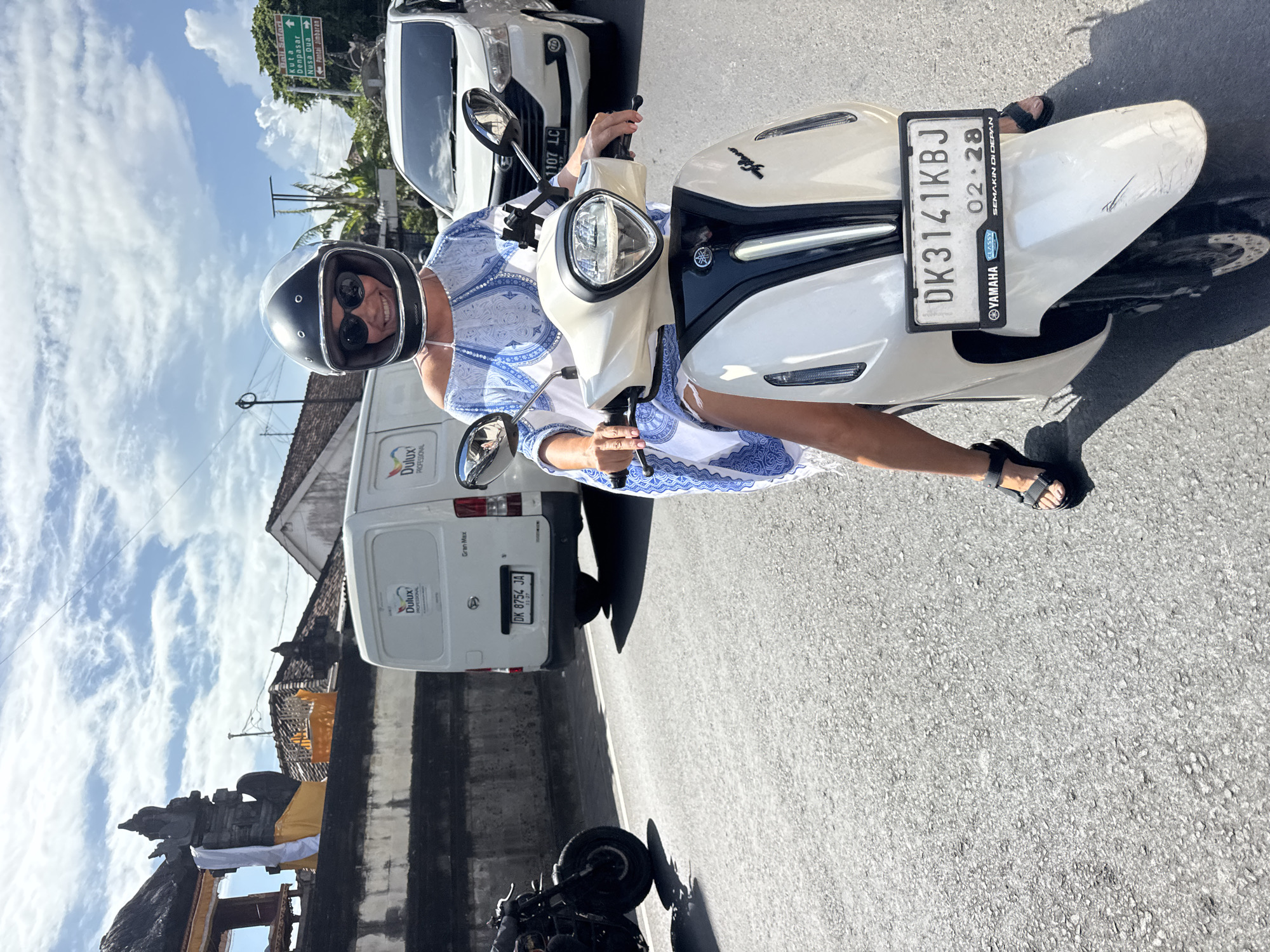 An overhead shot shows a woman wearing a black helmet and sunglasses, seated on a white Yamaha Classy scooter with the license plate DK3141KBJ, in Kuta Selatan, Indonesia. The scooter has a license plate sticker indicating a validation date of 02.28. She's wearing a blue and white patterned dress and black sandals.
A white van with "Dulux Professional" branding and license plate DK 8754 JA is visible to the left. Another white vehicle is partially visible behind the scooter. A sign indicating directions to "Bali Safari," "Kuta," "Denpasar," "Nusa Dua," and "Pantai Jimbaran" is in the background. Part of what seems to be a Balinese structure decorated with yellow cloth is visible to the left. The sky is partly cloudy.
