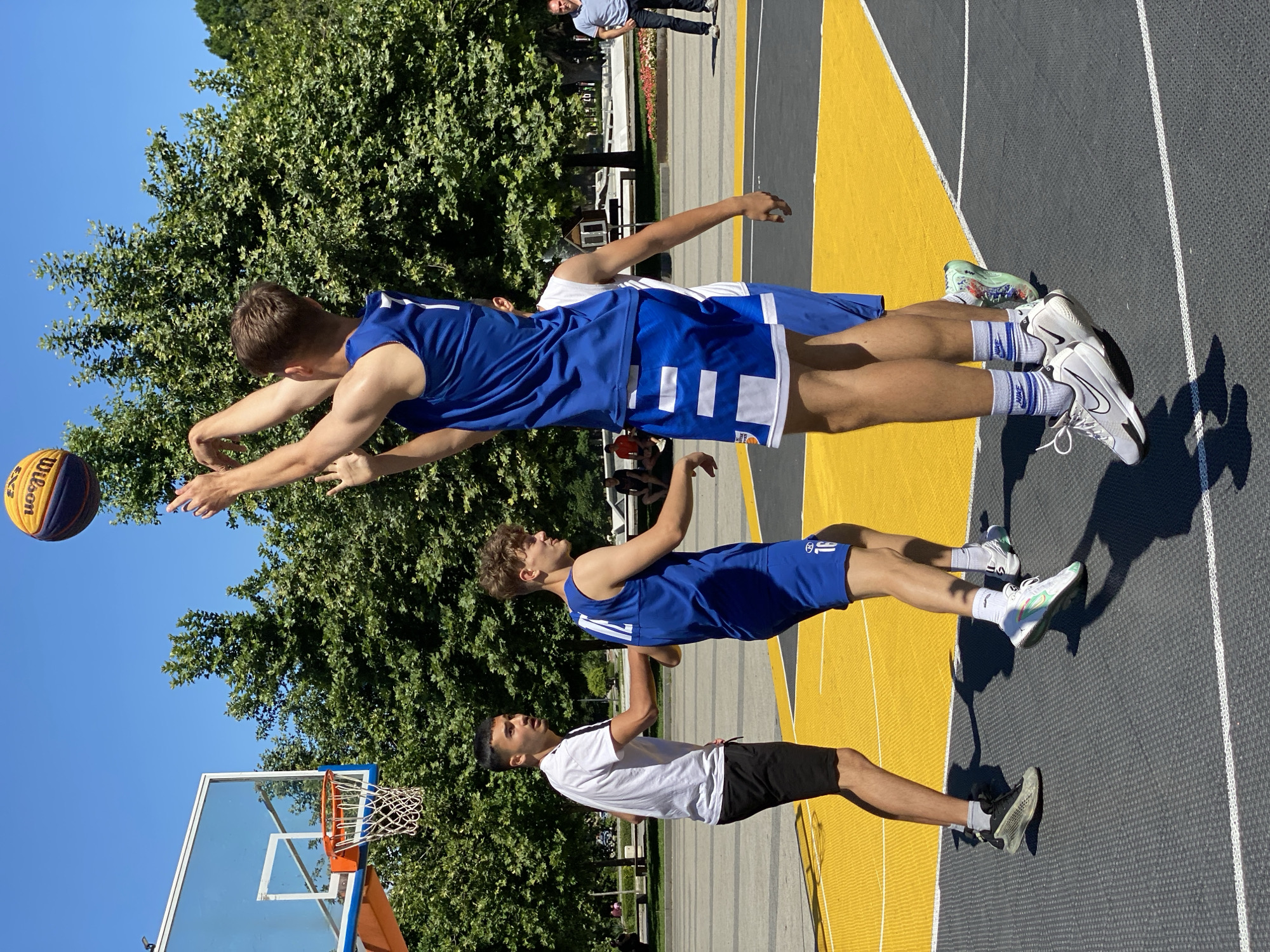 The image depicts an outdoor basketball court in Pleven, Bulgaria, with a clear blue sky and lush green trees in the background. Four young men are engaged in a game of basketball. In the foreground, a player in a blue jersey with the number 7 is mid-air, shooting a yellow and blue Wilson basketball. Another player in a blue jersey with the number 16 is positioned behind him, looking towards the basket. To the left, a player in a white t-shirt and black shorts is watching the play. A fourth player, mostly obscured, is in a white tank top and blue shorts. The court has distinct yellow and grey markings. In the background, several other individuals are visible, some sitting on benches and others standing. The overall scene captures a moment of athletic activity in a public park or recreational area in Pleven.