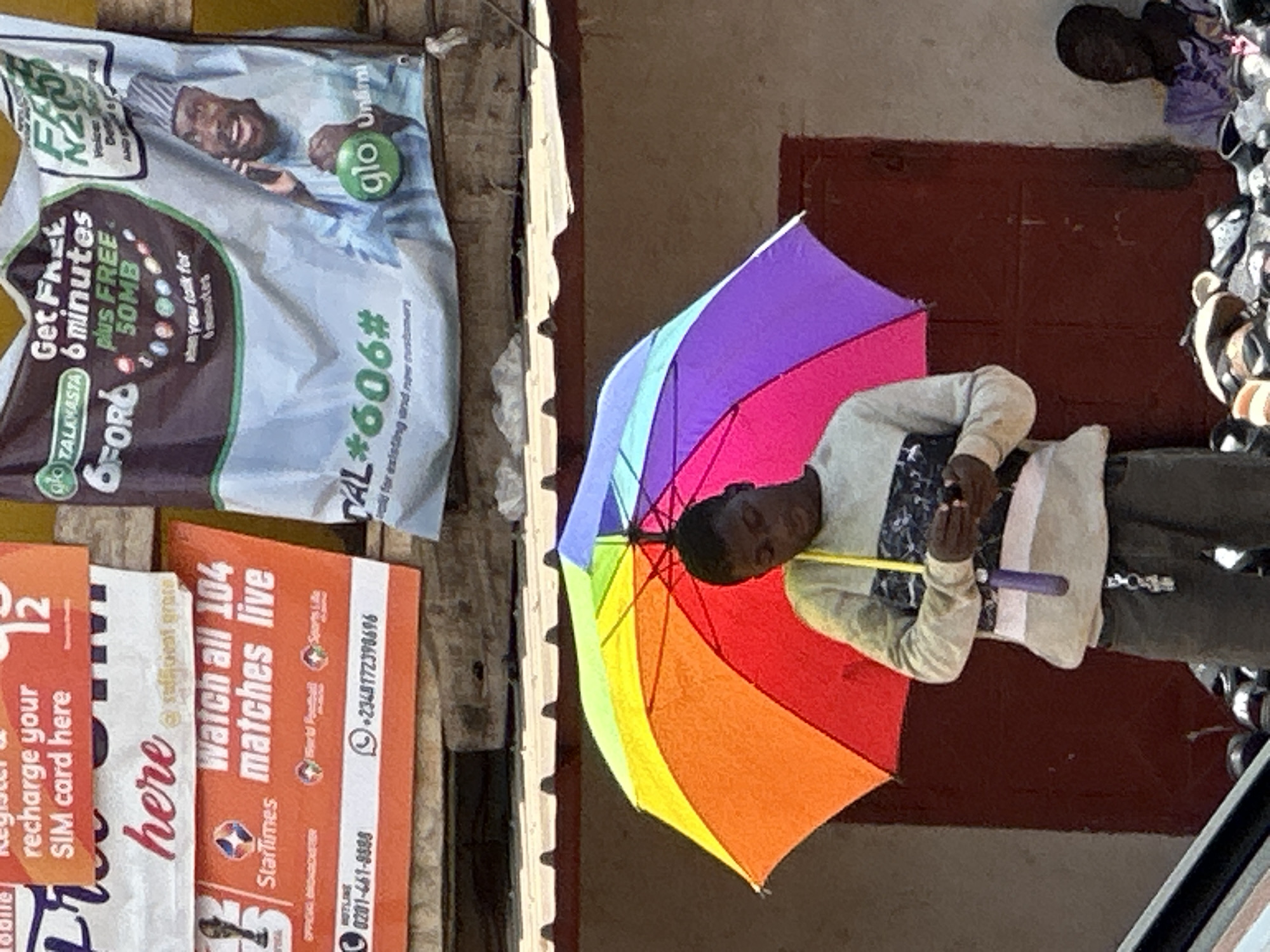 In the city of Bukuru, Nigeria, a young Black male stands beneath a rainbow-colored umbrella, holding a mobile phone. He is wearing a grey long-sleeved shirt with a patterned design across the chest and dark trousers. To his right, another Black male, wearing a purple shirt and a striped beanie, sits in front of a display of footwear. Above them, advertisements are visible, including one for Glo, a telecommunications company, offering free minutes and data. Another advertisement promotes watching live football matches. The scene suggests a bustling market or street-side vendor area.