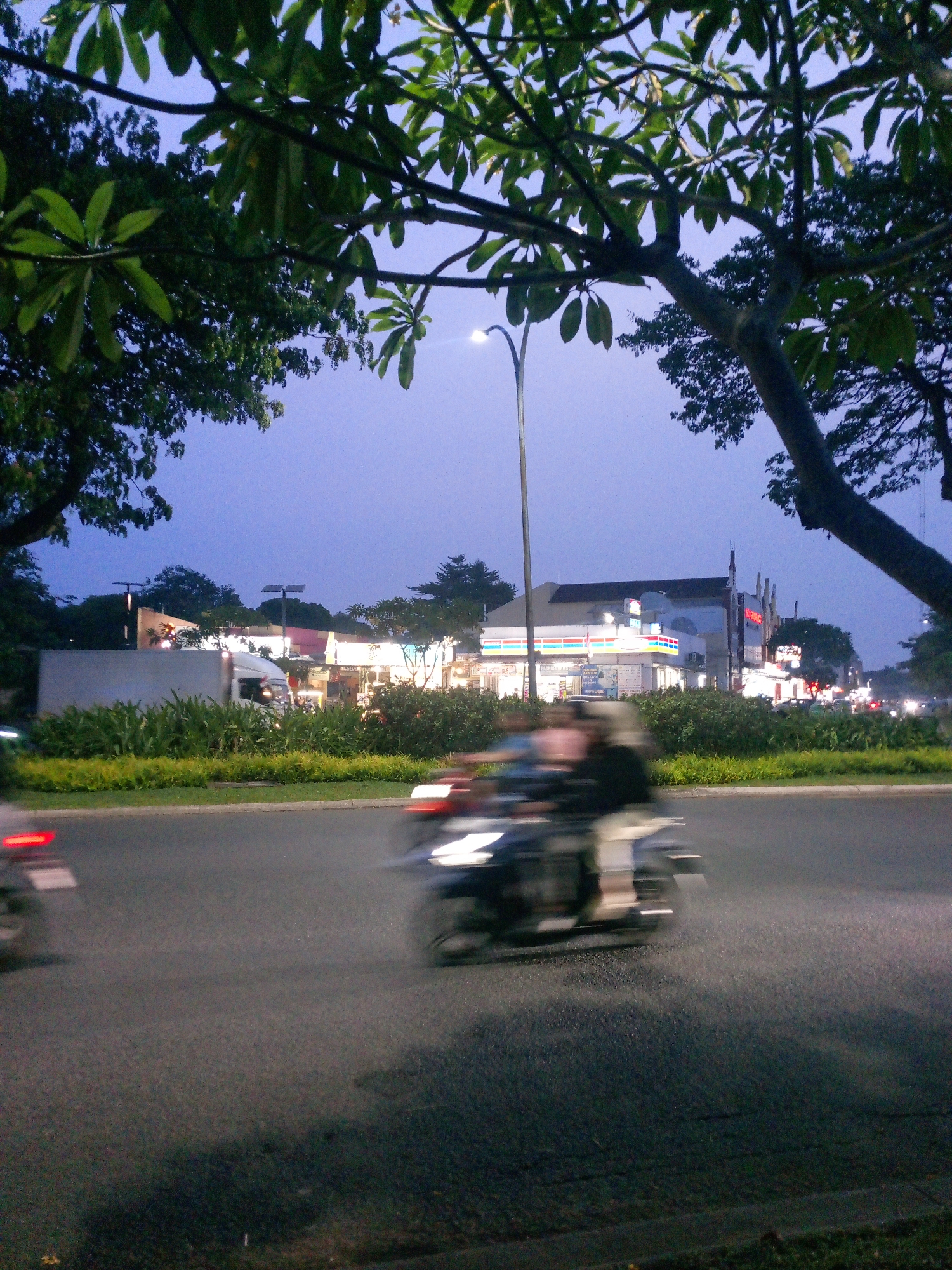 The image depicts a street scene at dusk in Cikupa, Indonesia. In the foreground, the asphalt road is visible with motion blur from passing vehicles, primarily motorcycles. One motorcycle in the center is captured with two people riding it, their forms blurred by movement. To the left, another motorcycle's red taillight is streaked across the road.
Behind the road, a median strip is covered with lush green bushes and yellow-green ornamental grass. Beyond this, a white truck is parked or moving slowly, with its headlights on. Further in the background, a row of buildings with illuminated signs suggests commercial establishments, possibly a convenience store and other shops, with their lights glowing against the twilight sky. Trees with dark green foliage frame the upper portion of the image, with some branches extending into the frame from the top. A tall streetlamp with its light on is positioned on the left side of the frame, extending upwards. The sky is a deep twilight blue.
