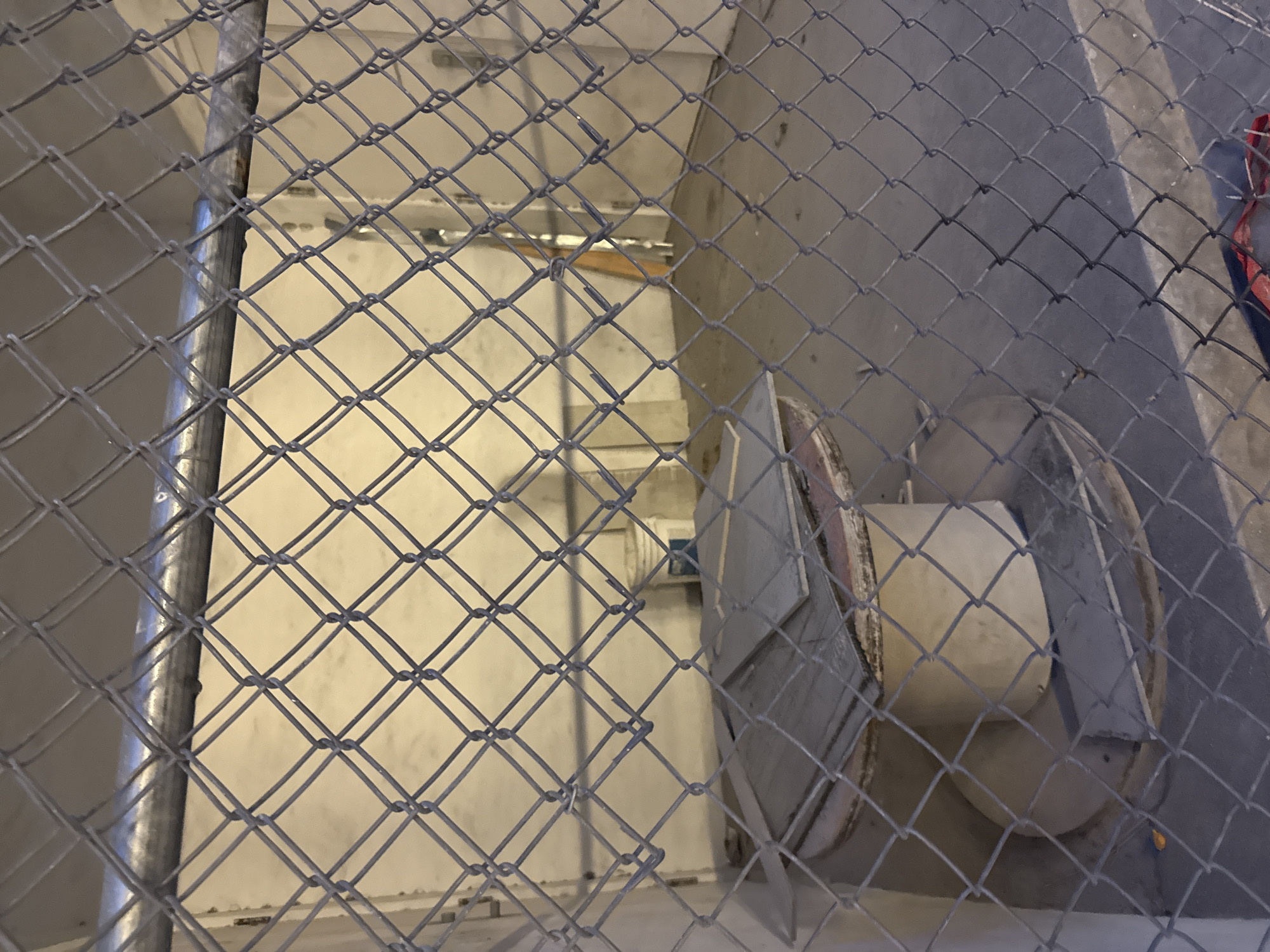 A high-angle, close-up shot through a chain-link fence shows an industrial or utility space. The area behind the fence appears to be a multi-level concrete structure with exposed pipes and supports. On the right side of the frame, behind the fence, there is a cylindrical object with metal housing. The exact location within the city of Miami, United States of America, cannot be determined from this image alone. No people are visible.