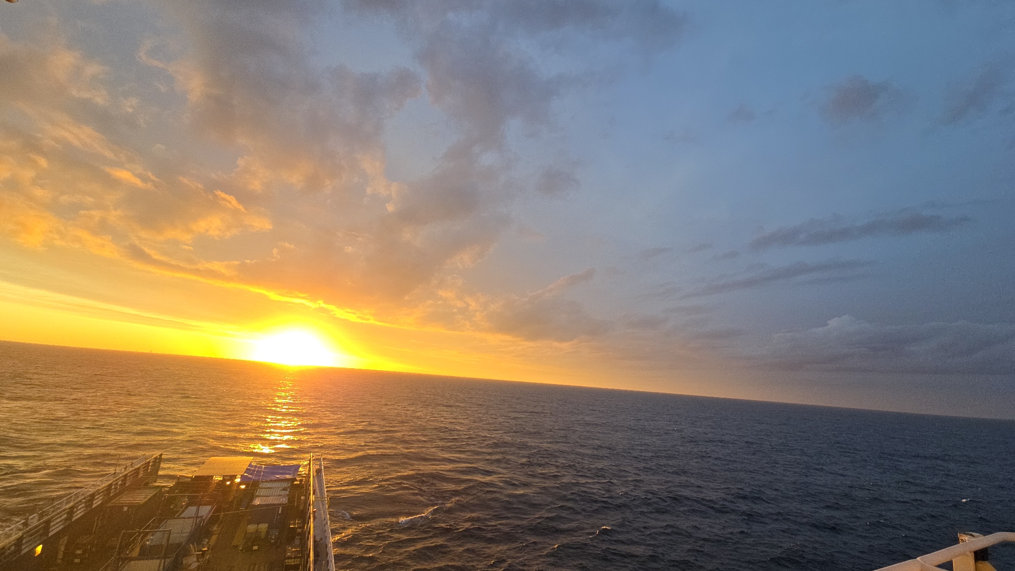 The image captures a sunset over the ocean from what appears to be a ship or vessel. The sun is low on the horizon, casting a bright, yellow-orange light across the water and the sky. The sky transitions from a fiery orange near the horizon to a dusky blue at the top of the frame, with some scattered clouds. The ocean is dark blue, with visible ripples and waves reflecting the sunlight. The foreground includes part of the ship's structure, showing metallic railings and what looks like cargo or equipment covered with tarps. The location and city are unknown.