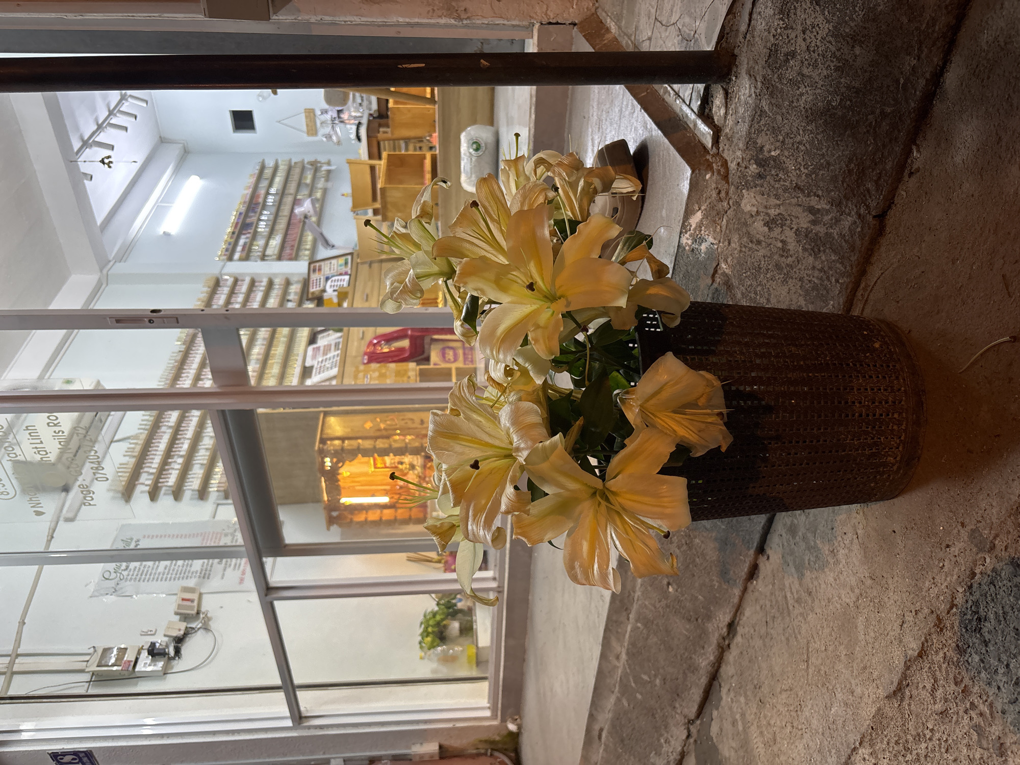 A bouquet of pale yellow lilies in a dark, woven cylindrical vase sits on a concrete sidewalk in front of a storefront. The storefront has large glass windows that reveal the interior. Inside, shelves are stocked with numerous small bottles, likely nail polish, and display cases hold nail art supplies. A desk and chairs are visible further inside, suggesting a nail salon. The exterior wall of the building is visible to the left, with electrical conduits and boxes mounted on it. The lighting suggests it is nighttime, with artificial lights illuminating the interior of the shop and casting a warm glow. The concrete ground is textured and appears wet in some areas, possibly from rain or cleaning. A metal pole stands to the right of the frame. A sign is visible through the window with text that includes "Nails Ro".