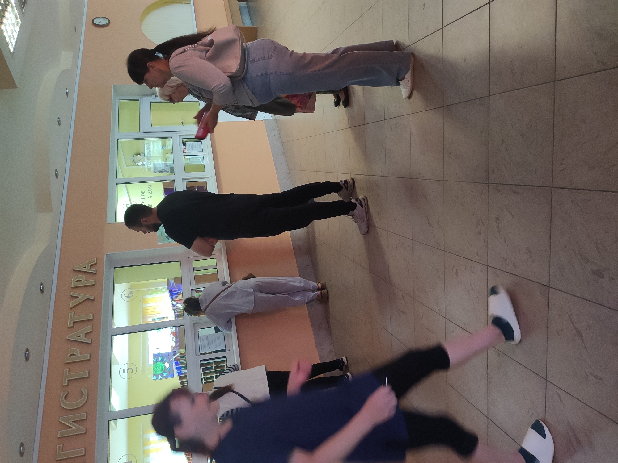 The image shows a registration desk with several people present. A woman in the foreground is walking towards the left, partially blurred due to motion. Behind her, a woman in a white top and dark pants stands at the counter. To the right of her, a person in a white uniform is visible. Further to the right, a man in a black t-shirt and dark pants is standing, facing the counter. A woman with a handbag and a man with a beard are also visible near the counter. The registration desk has windows with numbers 5 and 6 displayed. A clock is mounted on the wall to the right. The environment appears to be an indoor public building, likely a government institution or clinic. The text above the windows reads "ГИСTРАТУРА" (REGISTRATURA), suggesting a registration office. The overall scene depicts a typical waiting or service area. Without specific knowledge of Navapolatsk, Belarus, it is impossible to determine the exact location within the city, however, the overall context suggests a public service building.