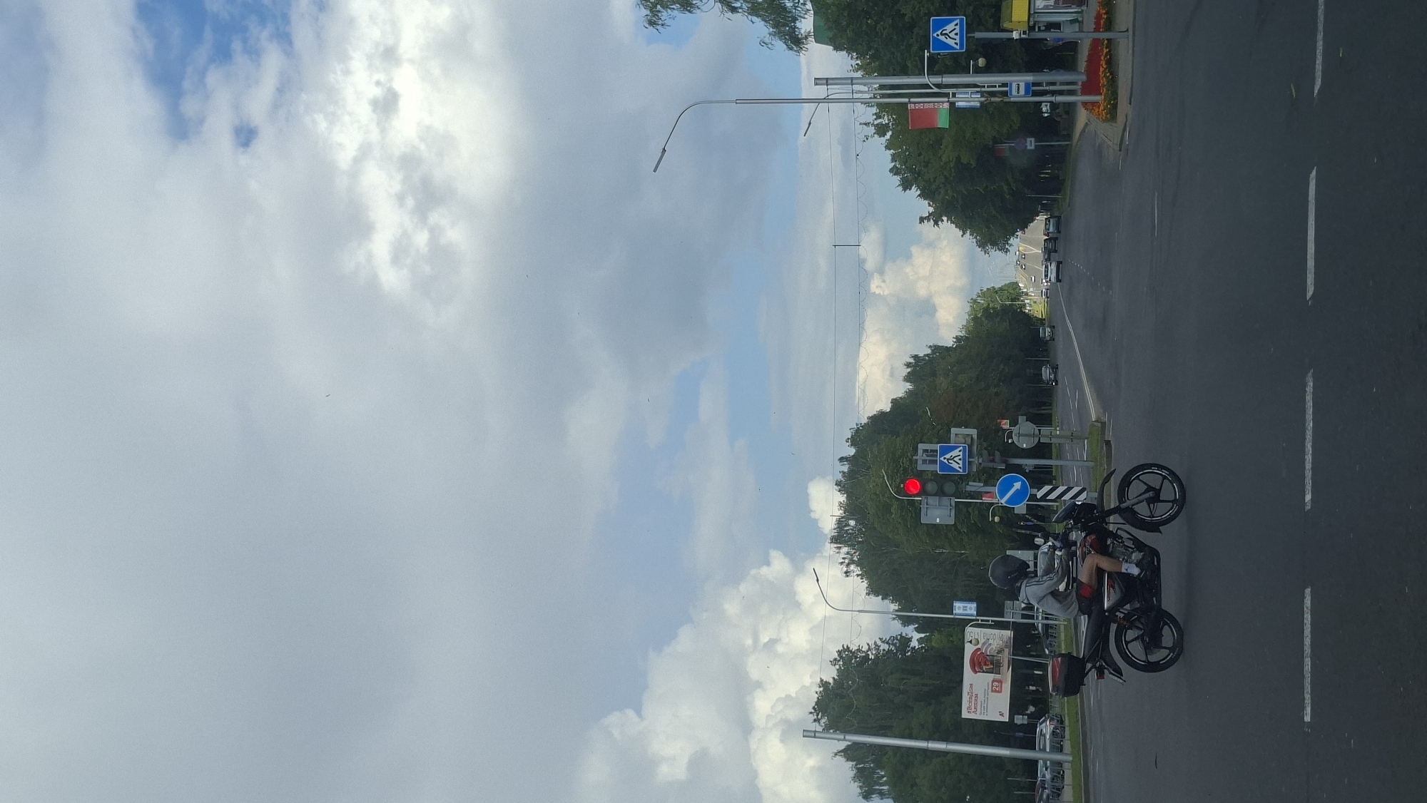 This image depicts a street scene in Maladzyechna, Belarus, on a cloudy day. The focal point is a motorcyclist wearing a grey hooded sweatshirt, red shorts, and a black helmet, riding a black motorcycle with a red top case. The motorcycle is moving across an intersection. In the background, there are traffic lights, pedestrian crossing signs, and various trees lining the street. Several cars are visible on the road in the distance. The sky is filled with large, dramatic clouds. A billboard for "Vesba" is visible on the left side of the image.