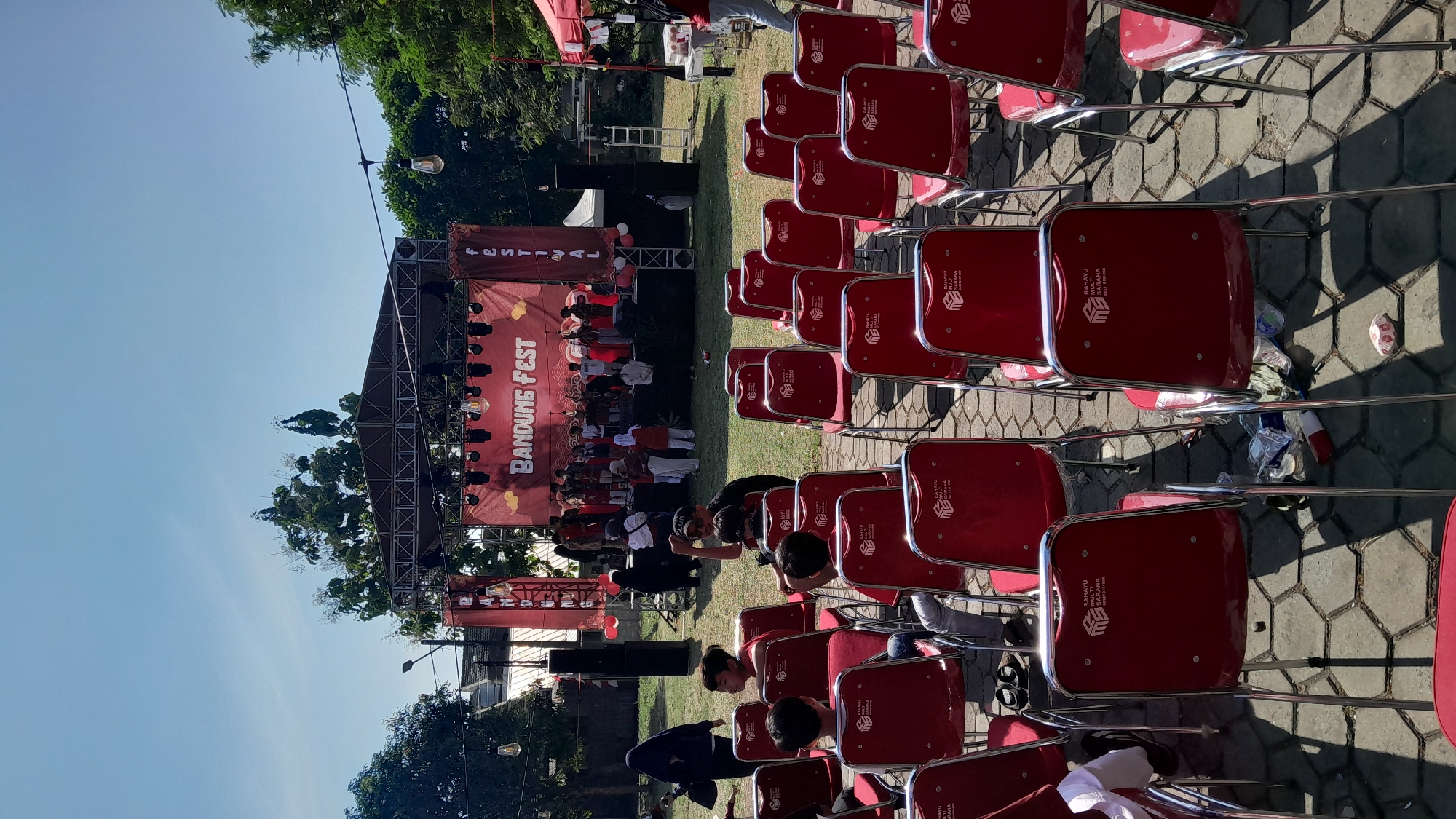 An outdoor festival stage with a banner reading "BANDUNG FEST" is set up for a performance. A group of performers, some in uniform, are on stage. In front of the stage, rows of empty red chairs are arranged on a paved area. Several people are standing in the background, including some wearing hijabs. Trees and a clear blue sky are visible in the background. The lighting suggests it is daytime.