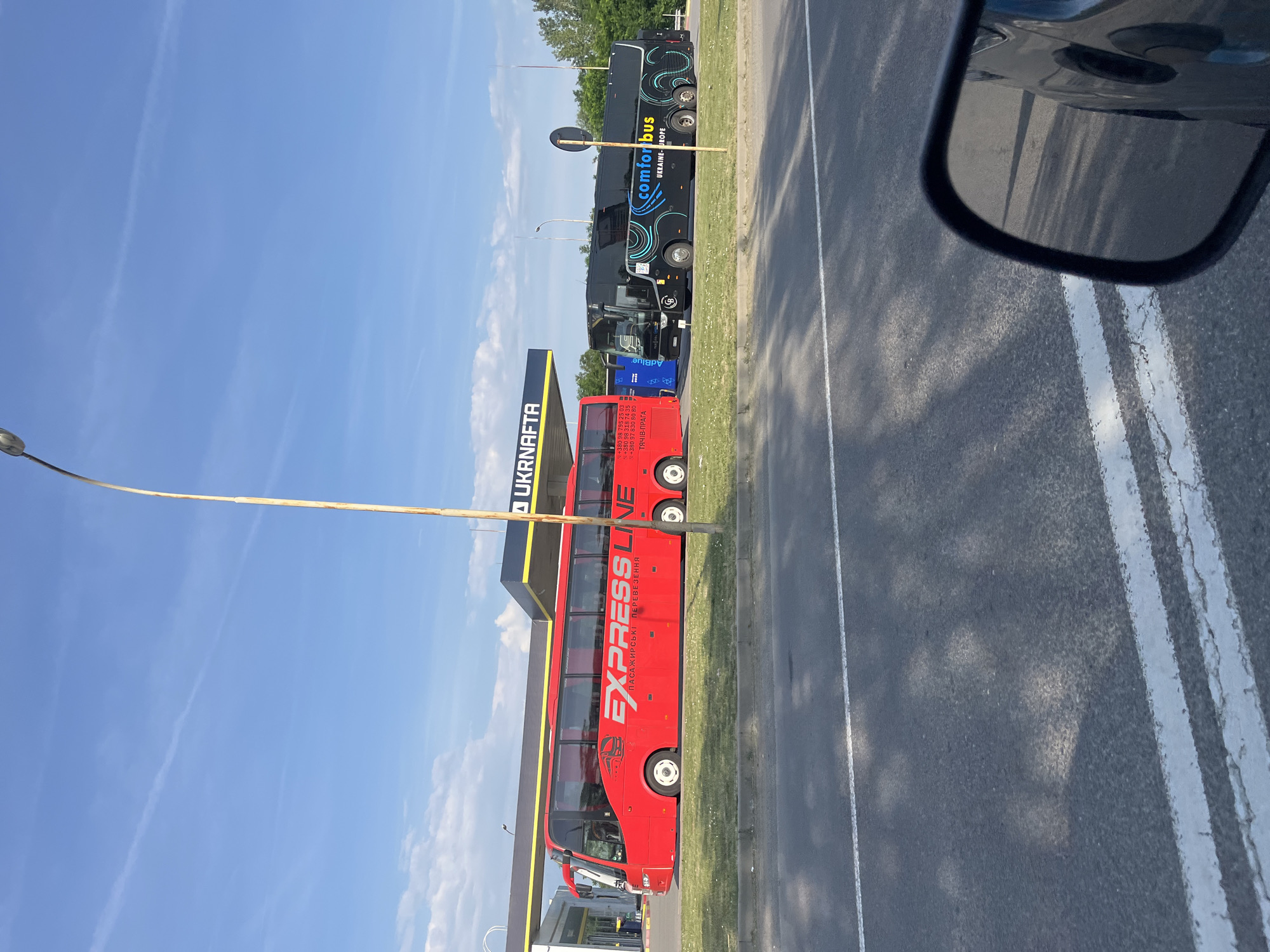 In Baranyntsi, Ukraine, two tour buses are parked under a Ukrnafta gas station canopy. The bus on the left is red and has "EXPRESS LINE" written on its side in white lettering. The bus on the right is black and features "comfort bus UKRAINE EUROPE" in colorful lettering. The scene is captured from a low angle, showing the road in the foreground and the buses parked on a grassy area with the gas station behind them. The sky is blue with some clouds.