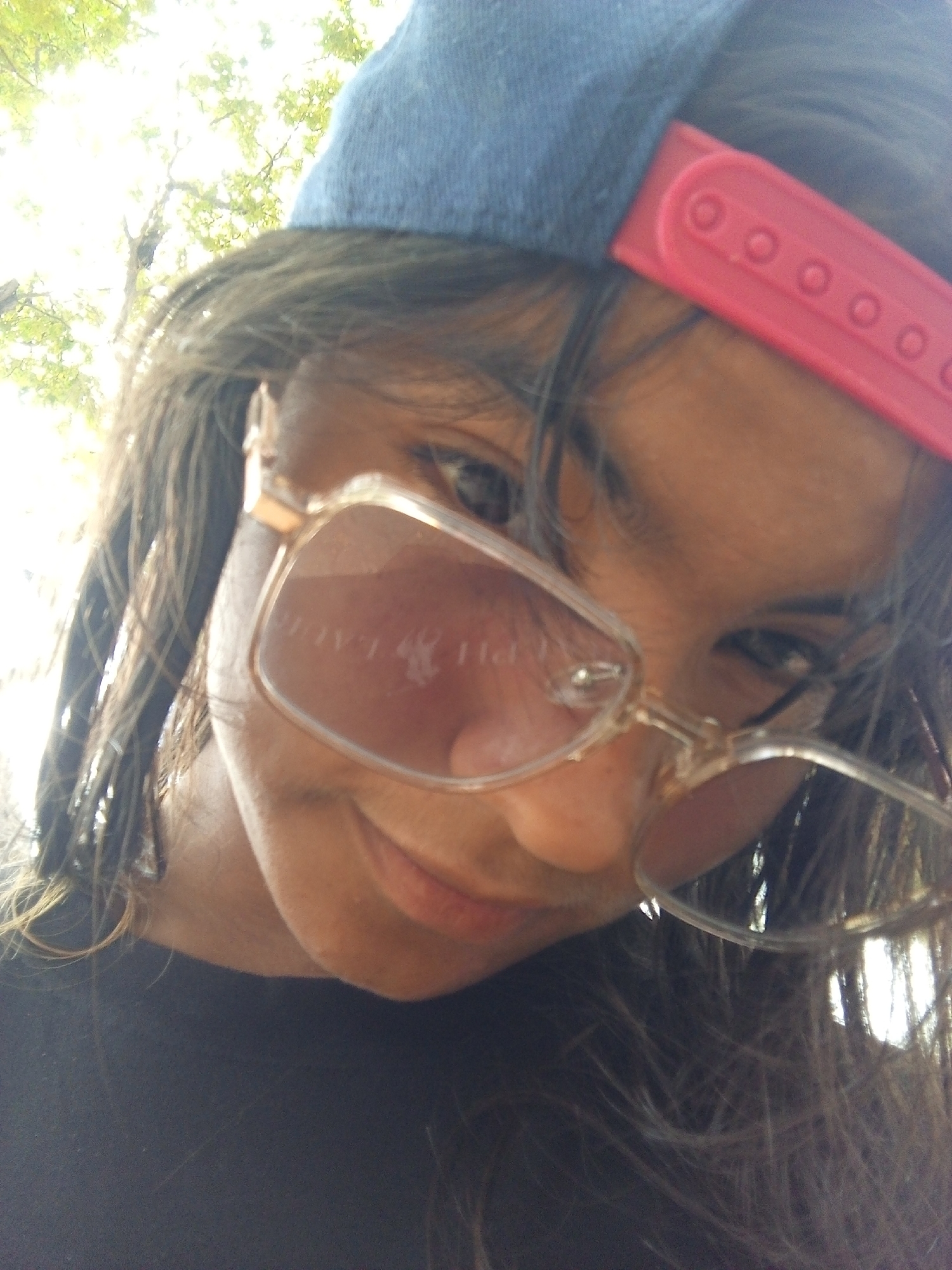 A close-up, low-angle shot shows a person with dark hair, wearing transparent-framed eyeglasses, a blue baseball cap, and a black shirt. The baseball cap has a pink adjustable strap. The eyeglasses appear to have a mirrored reflection. The person is looking down and slightly to the side. The background is blurred, showing green foliage and sunlight. The location is unknown, and there is no verifiable evidence in the image that identifies Juan Griego, Venezuela, as the location.
