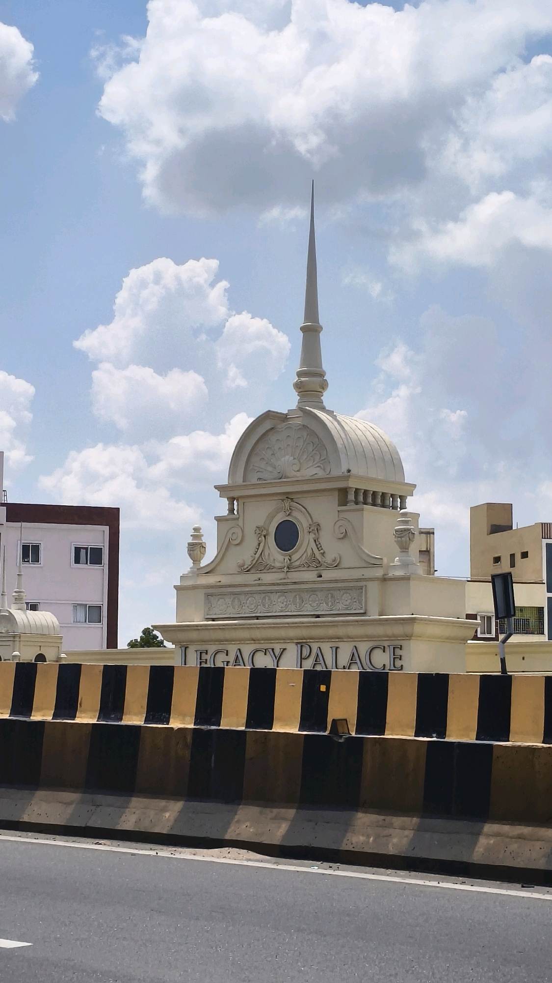 The image depicts the "Legacy Palace" building, a cream-colored structure with a dome and a spire, located in Hyderabad, India. The building features ornate architectural details, including decorative carvings, a circular blue window, and urn-like ornaments. The name "LEGACY PALACE" is prominently displayed in dark lettering on the building's facade.
In the foreground, a concrete road divider with alternating black and yellow stripes runs horizontally across the image, suggesting the viewpoint is from a road. Behind the road divider, another section of the building is visible, featuring a simpler design with rectangular windows. To the left of the Legacy Palace, a multi-story building with a brown and white exterior and several windows is partially visible. To the right, another building with a light brown facade and some windows is also in view.
The sky above is a bright blue with scattered white clouds. The overall scene appears to be an urban environment on a clear day.