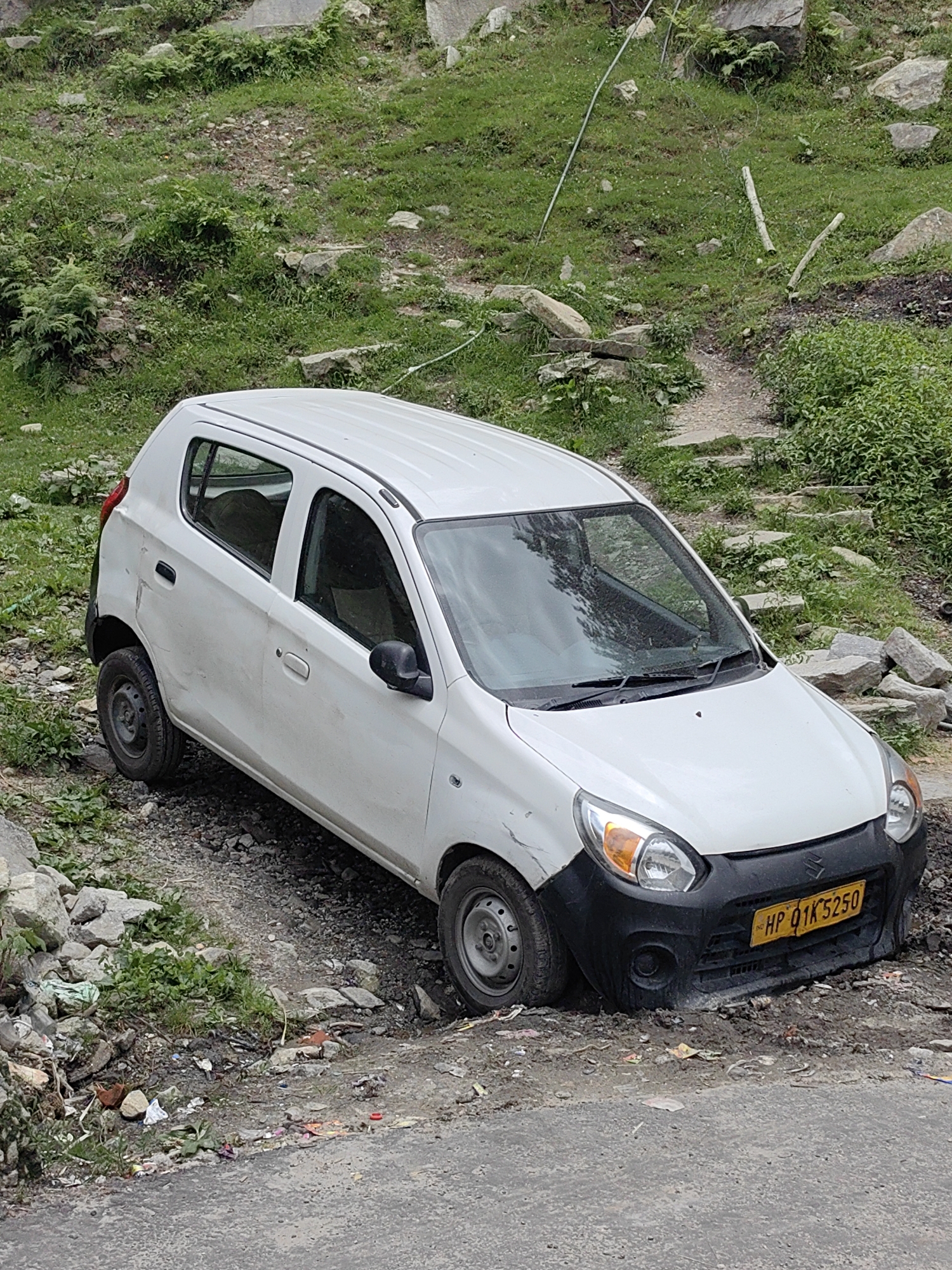 In Vashisht, India, a white Maruti Suzuki Alto 800 car, bearing the license plate HP 01K 5250, is partially embedded in a muddy embankment. The car is positioned at an angle, with its front right wheel deeply sunken into the soil and debris. The surrounding environment consists of a rough, unpaved surface with loose stones, rocks, and scattered trash. Green vegetation and grass are visible on the hilly terrain behind the car, and a rough path with stone steps leads up the slope. The car's front bumper appears to be black, and there are minor dents and scratches visible on its white bodywork. The scene suggests the car may have gone off the road or become stuck in the difficult terrain. There are no people visible in the image.