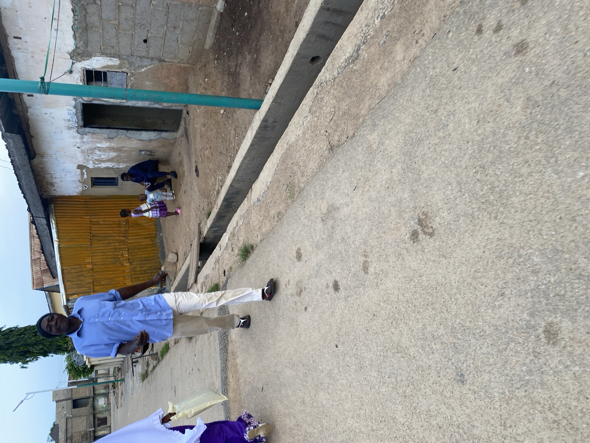 In Jos, Nigeria, a man in a light blue collared shirt and beige pants walks along a paved street, looking at the camera. He wears a dark beanie and sandals. To his left, another person in a purple garment walks away from the camera, carrying a light-colored bag. In the background, a man sits on a step outside a weathered building, watching two young children. The building has a textured, off-white facade with a dark doorway and a small window. A corrugated yellow wall stands behind the children. A drainage ditch runs alongside the street in the foreground. The sky is overcast.