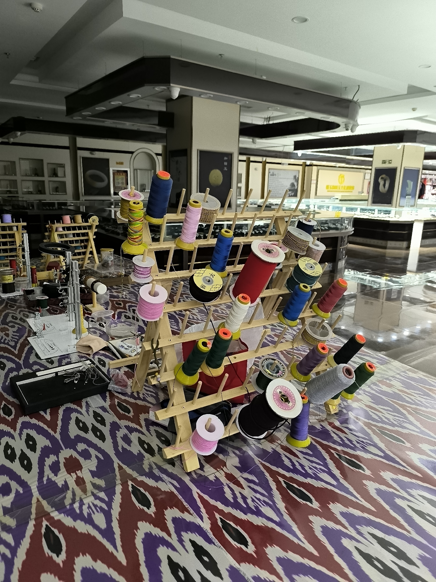 The image shows a display of spools of thread on wooden racks, set up on a patterned surface that appears to be a table or a display counter. The spools are of various colors including blue, pink, yellow, red, green, and black. Some spools are larger than others. In the background, there are display cases that suggest a retail environment, possibly a jewelry store or a craft supply store. The lighting in the background is artificial, and the overall impression is that of an indoor commercial setting. The patterned surface in the foreground is a complex design of purple, red, black, and off-white shapes. This display of threads is likely in Ürümqi, China, based on the request.