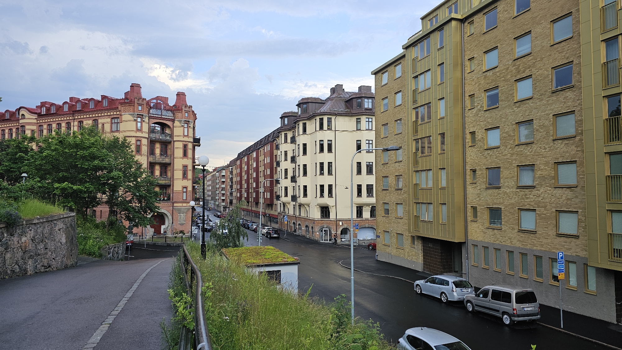 The image depicts a view of a street in Gothenburg, Sweden, characterized by multi-story apartment buildings and a winding road. The perspective is from an elevated position, looking down a curved asphalt road that slopes gently downwards. On the left, a substantial, ornate building with a red tiled roof and yellow brick facade is prominent. This building features balconies and numerous windows. Adjacent to it, a stone retaining wall supports a grassy embankment with lush green vegetation.
To the right of the road, a series of modern apartment buildings line the street. The closest building on the right is constructed with yellow brick and features a metallic gold-colored facade cladding. This building has multiple floors with regularly spaced windows and balconies. Further down the street, other apartment buildings of varying heights and architectural styles are visible, including one with a red facade and another with a light beige facade.
Parked cars are present along the street, including silver sedans and a gray van. A small, modern structure with a green vegetated roof is situated on the side of the road, possibly a utility building or information kiosk. Streetlights are visible, illuminating the scene. The sky is overcast with scattered clouds. The overall impression is of a residential urban environment with a mix of older and newer architecture.