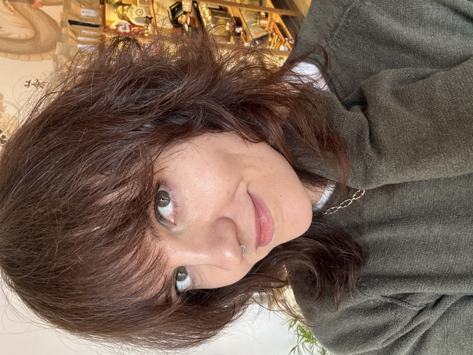 A female subject, approximately 20-40 years old, with dark brown, shoulder-length, layered hair and bangs, and hazel eyes, is depicted in a frontal head-and-shoulders portrait. Her expression is a slight smile, and she looks directly at the camera. She has a silver septum ring and wears a silver-colored chain necklace over an olive green crew-neck sweater, layered above a white undergarment. The background reveals an indoor retail setting. To the subject's left, a white wall and green foliage are visible. To the right, a light-toned wall features an East Asian-style mural with stylized designs and characters. Below the mural, multiple shelves display various items, including ornate gold-colored vessels, dark figurines, and small packaged goods, consistent with a specialty tea or gift shop. While the specific street or establishment name within Budva, Montenegro, is not identifiable, the interior design suggests a commercial space typical of tourist-oriented shops.