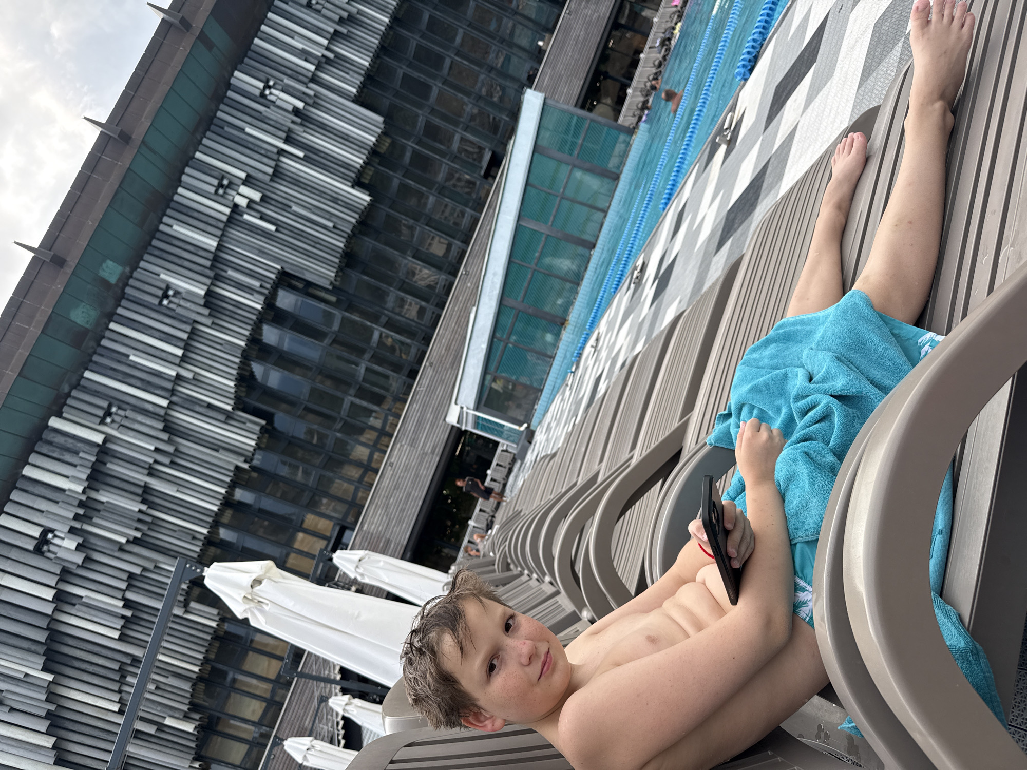 The image shows a young boy lying on a lounge chair near a swimming pool. He is wearing swim trunks and has a blue towel draped over him. The boy is holding a black smartphone in his right hand and appears to be looking at the camera with a slight smile. His hair is wet, suggesting he has recently been in the water.
The setting is a modern outdoor space with a swimming pool in the background, marked by blue lane dividers. Deck chairs line the pool area, and a contemporary building with a distinctive facade featuring horizontal elements is visible behind them. White umbrellas are also set up in the area, suggesting it's a place for relaxation and leisure.
The lighting suggests it could be daytime, possibly late afternoon given the angle of the light. The sky is overcast. The overall mood is relaxed and casual, consistent with a vacation or pool day.
No visible text is present in the image. The location context is provided as the city of Yalta, Ukraine.