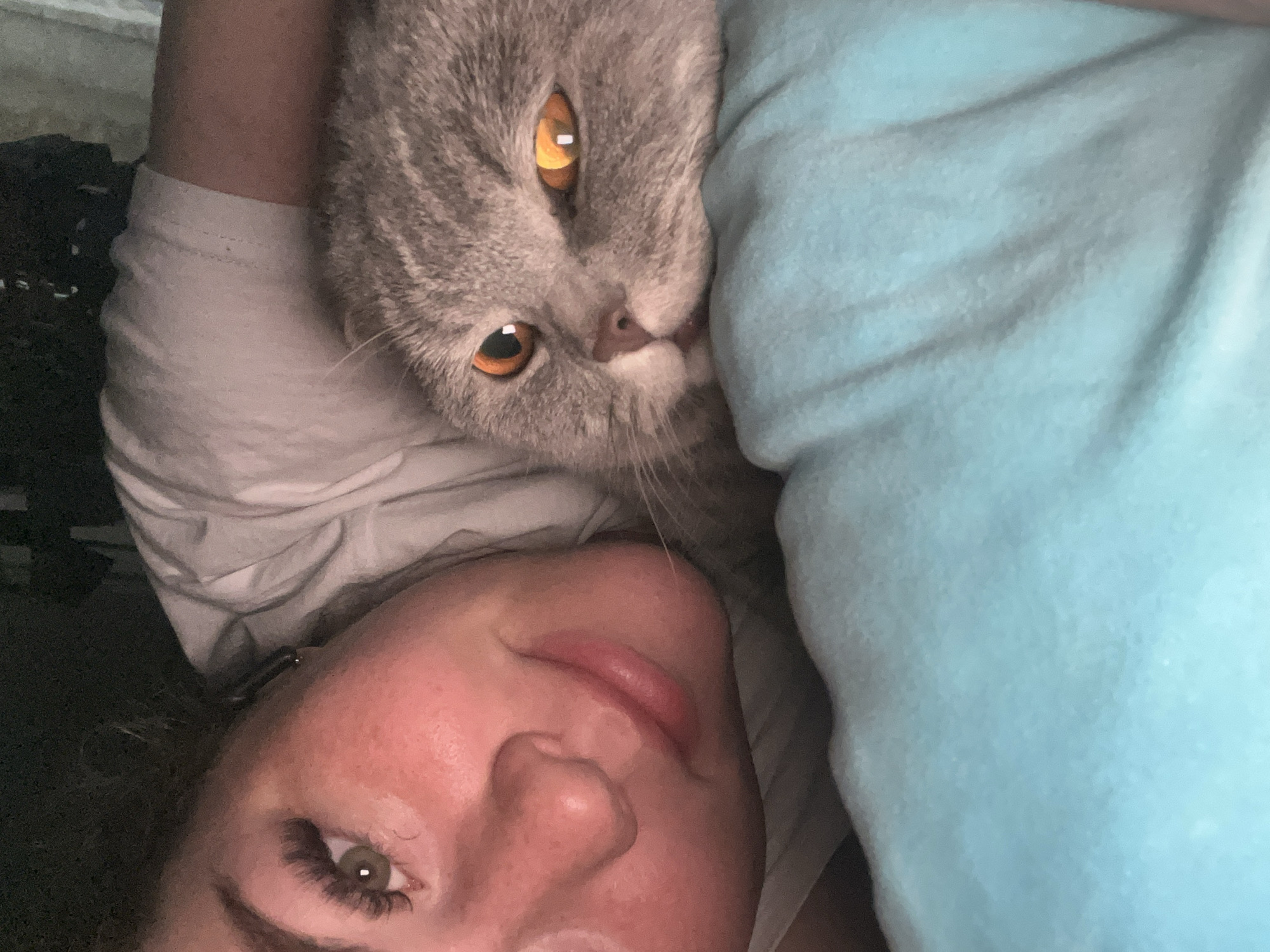 A woman lies on her back with her head tilted slightly towards the bottom of the frame. A gray cat rests its head on her shoulder, its amber eyes gazing upward. The woman has fair skin, green eyes, and long eyelashes. A light blue pillow is positioned next to her head. The background is indistinct, revealing glimpses of a white t-shirt and some dark-colored objects. The location in the city of Chaikovsky, Russia, is not determinable from this image.