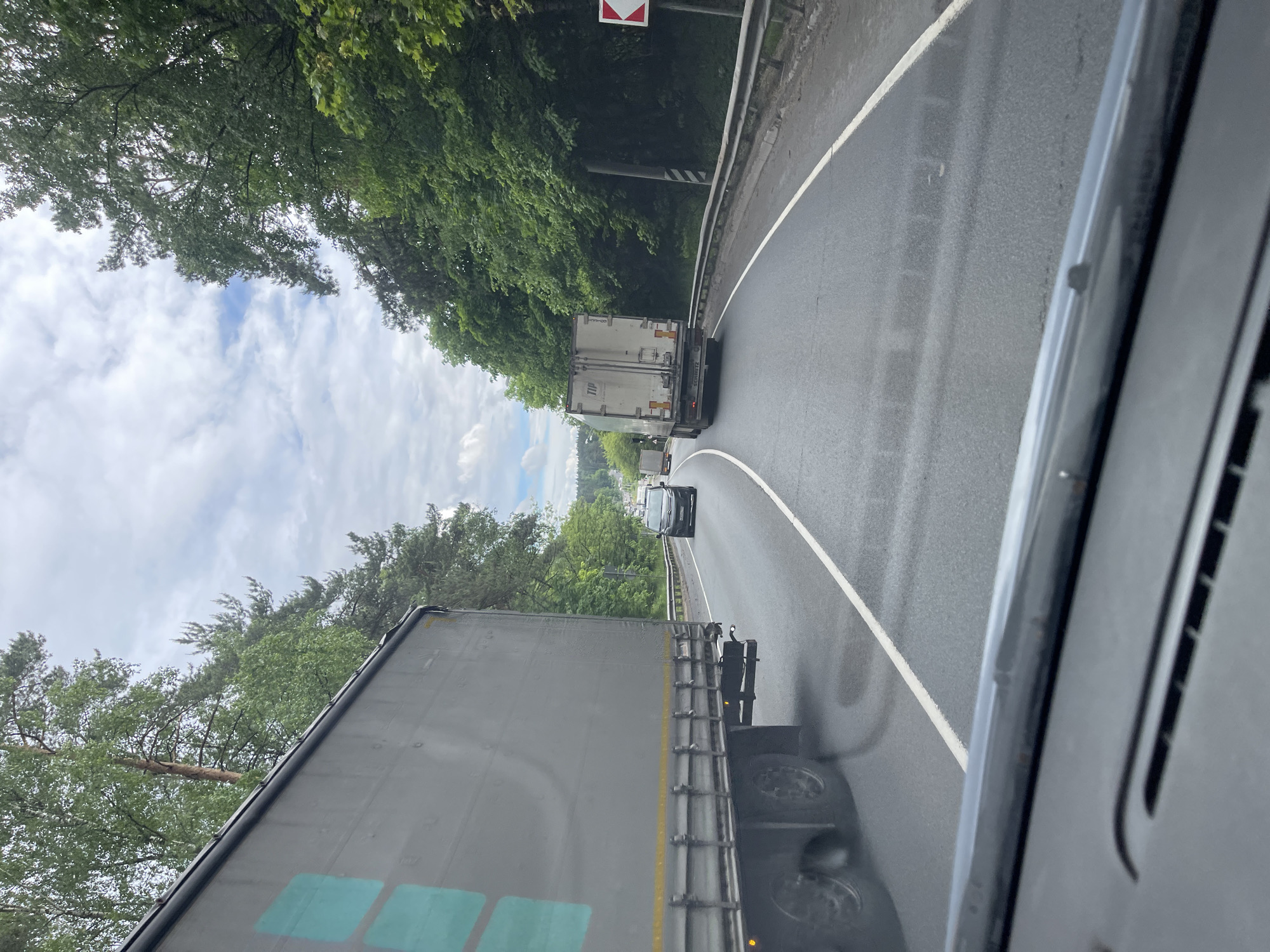The image shows a view from inside a vehicle, likely a truck, traveling on a wet, winding road. In the foreground, a large truck is visible, taking up much of the lower half of the frame. Ahead of the truck, a black car is following another truck. The road curves to the right and is bordered by a metal guardrail. Beyond the guardrail, dense green trees line the road. A road sign is visible on the right side of the road. The sky is overcast with patches of blue visible through the trees. It is not possible to confirm the location within Semyonovskoye, Russia without additional information.