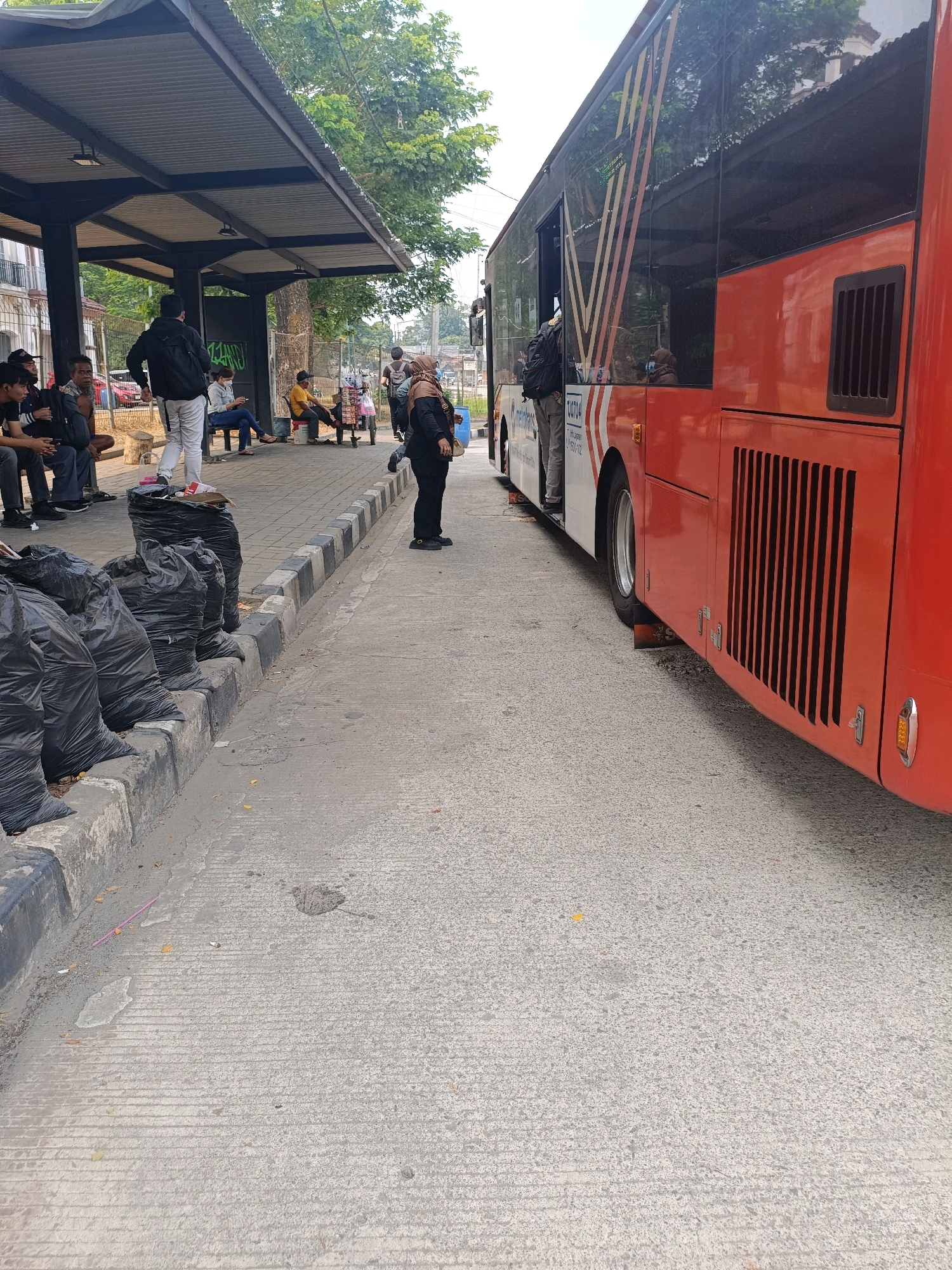 The image depicts a bus stop in Tangerang, Indonesia, with a red and white bus parked alongside the platform. Several people are waiting for the bus, some seated on the bench and others standing. Black trash bags are piled up near the curb. The bus stop shelter is visible in the background, with trees and buildings beyond. The overall scene suggests a typical urban bus stop environment.