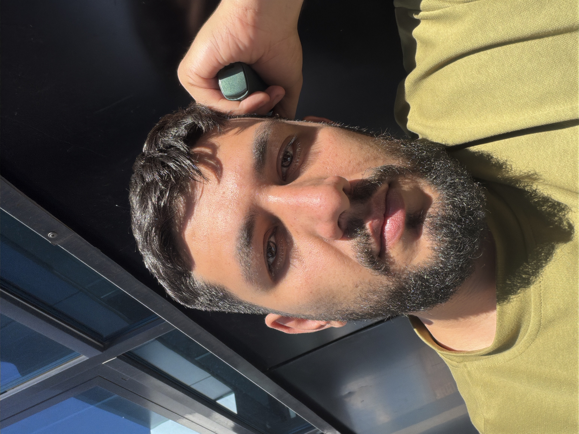 A man with dark hair and a beard is holding a small, green electronic device to his head in an outdoor setting. He is wearing a light green shirt. Sunlight is casting shadows on his face and shirt. Behind him, there is a black surface and a window with a view of the sky. The location cannot be determined definitively from this image, and the city of Krasnoyarsk, Russia is not able to be confirmed from the image.