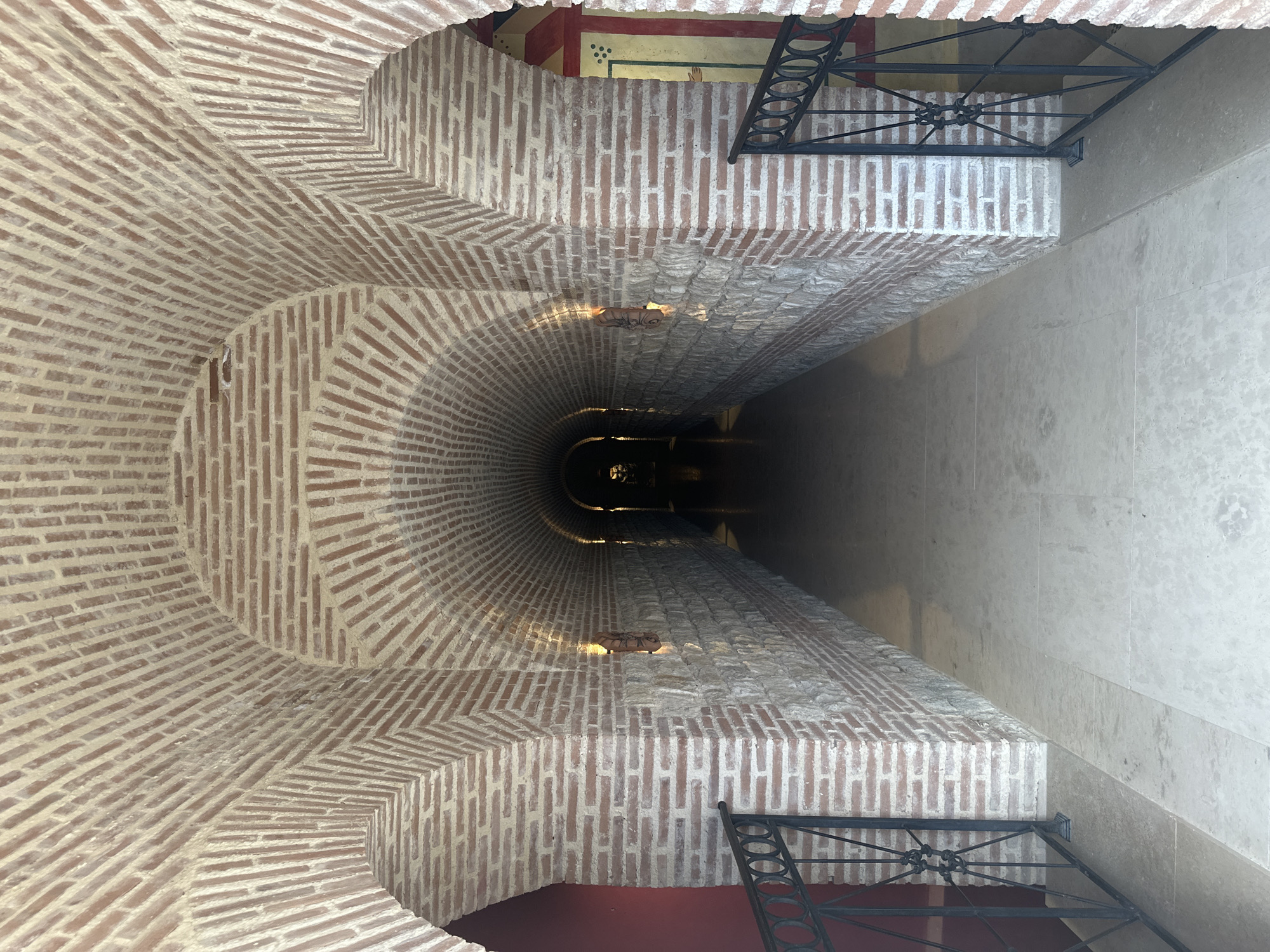 The image shows a view down a brick arched tunnel. The walls are constructed from alternating layers of red brick and stone, with decorative lighting fixtures mounted on the stone sections. The tunnel has a paved floor and extends into darkness at the far end, where a dimly lit area is visible. Black metal railings with decorative elements are present on either side of the tunnel's entrance. The scene appears to be an architectural interior.