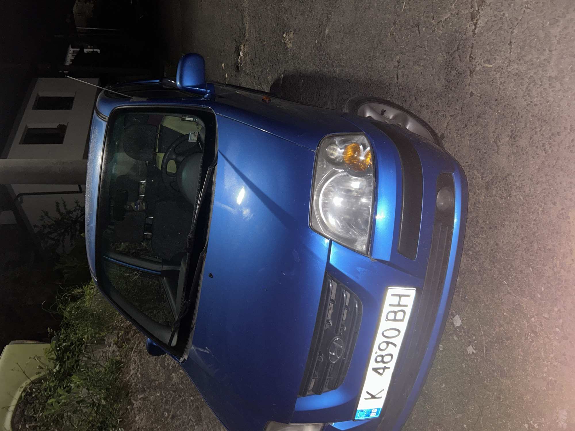 A blue car, with a Bulgarian license plate that reads "K 4890 BH," is parked on the side of what appears to be a paved road. The car is facing towards the viewer's right. A building is visible in the background on the left side of the image. The setting is nighttime. The location might be in the city of Pripek, Bulgaria.