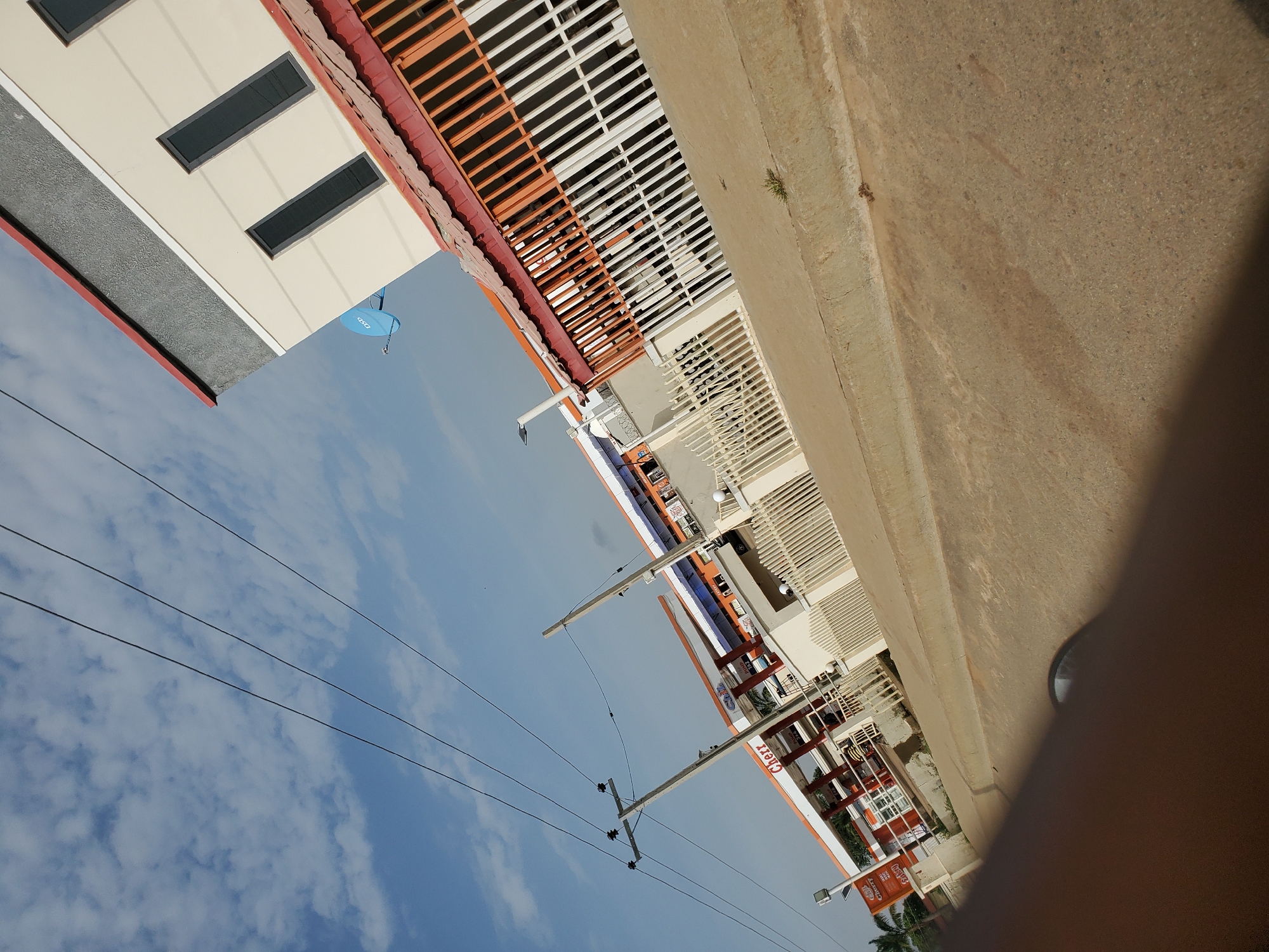 The image depicts a street scene in Bukuru, Nigeria, featuring a Cherr gas station. The sky is partially cloudy with a bright blue expanse. Overhead power lines traverse the frame diagonally. The Cherr gas station has an orange and white canopy covering the pumps, and a white fenced perimeter. To the right, a modern building with a gray and cream facade and tall, dark-tinted windows is visible, topped with a red roof. A blue satellite dish is mounted on the side of this building. The road in the foreground is paved and appears to be asphalt. A portion of a vehicle's interior, likely the dashboard and windshield area, is visible in the bottom left corner of the frame, suggesting the image was taken from inside a moving vehicle.