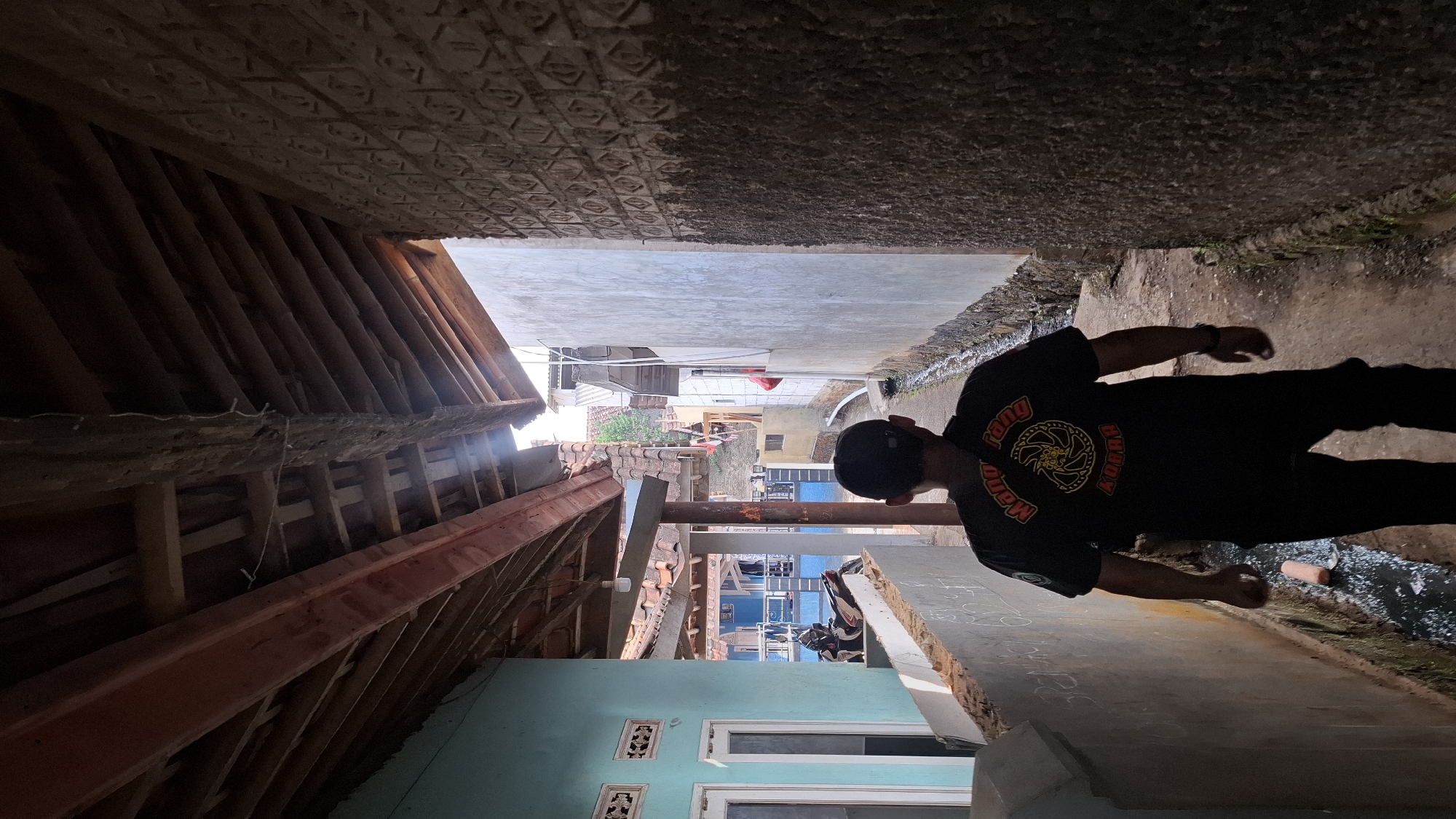 The image shows a person walking down a narrow alleyway. The person is wearing a black t-shirt with a yellow emblem and a black baseball cap. The alleyway is lined with buildings on both sides. On the left, there is a light blue building with windows and a motorcycle parked in front. On the right, there is a wall with a textured pattern. The alleyway has a dirt path and a drainage ditch running alongside it. The overhead view reveals wooden beams and roof tiles. The scene is likely in a residential area of Cilaku, Indonesia, given the architectural style and the presence of a motorcycle, common in Indonesian urban or suburban settings. The lighting suggests it is daytime, with sunlight filtering through the gaps in the roofing structures.