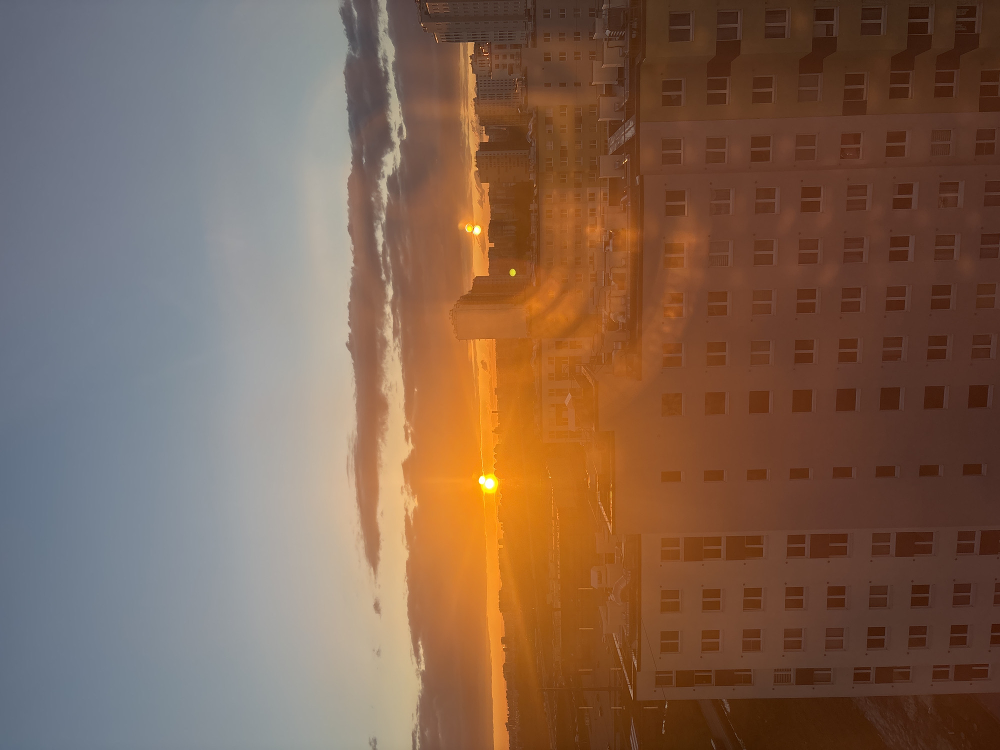The image captures a sunrise or sunset scene in Vsevolozhsk, Russia, with an intense golden light illuminating the cityscape. Tall apartment buildings dominate the right side of the frame, their facades punctuated by numerous windows. The sun, appearing as a bright orb, is partially obscured by clouds, creating a dramatic sky. The horizon is filled with buildings and other urban elements. The image is rotated 90 degrees clockwise.