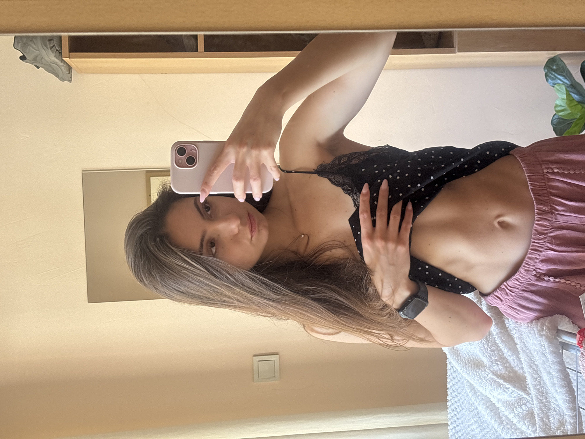 The image is a selfie taken in a mirror, featuring a young woman with long, blonde hair. She is wearing a black, polka-dotted top with lace trim and pink, ruffled shorts. Her left hand is holding a pink smartphone, and her right hand is resting on her stomach. A black smartwatch is visible on her left wrist. In the reflection of the mirror, a shelf with a shoe is visible. To the right of the woman, a green plant with large leaves can be seen. The overall scene appears to be indoors, possibly a bedroom or bathroom, given the presence of a mirror and a white towel or rug. The lighting suggests daytime. The woman's expression is neutral, and she is looking directly at the camera. The location is Varna, Bulgaria.