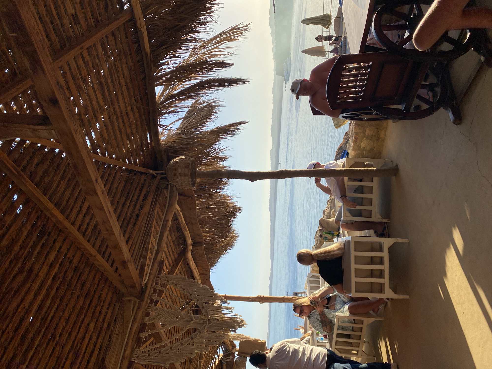 The image depicts a seaside restaurant or cafe in Sharm El Sheikh, Egypt, likely during the late afternoon or early evening, given the warm, low sunlight casting long shadows. The establishment features a rustic, open-air design with a thatched roof supported by wooden beams. Palm fronds hang down, adding to the tropical ambiance.
Several people are seated at tables, enjoying the view of the calm blue sea and the distant, hazy hills. In the foreground, a man wearing a white baseball cap and no shirt is seated in a distinctive wooden chair designed to resemble a wagon wheel. To his left, a woman with blonde hair in a ponytail is sitting at a white outdoor sofa, holding her phone. Further to the left, another woman with blonde hair and sunglasses is seated, also holding a phone. A man in a light-colored shirt is standing on the far left, facing away from the camera.
In the background, the sea stretches out, with a few boats visible in the distance. Umbrellas are set up on what appears to be a paved area near the tables, suggesting a lounging or dining space. The overall atmosphere is relaxed and leisurely, indicative of a vacation setting. The architecture and the presence of the sea strongly suggest a coastal location in Sharm El Sheikh, known for its resort amenities and beachfront establishments.