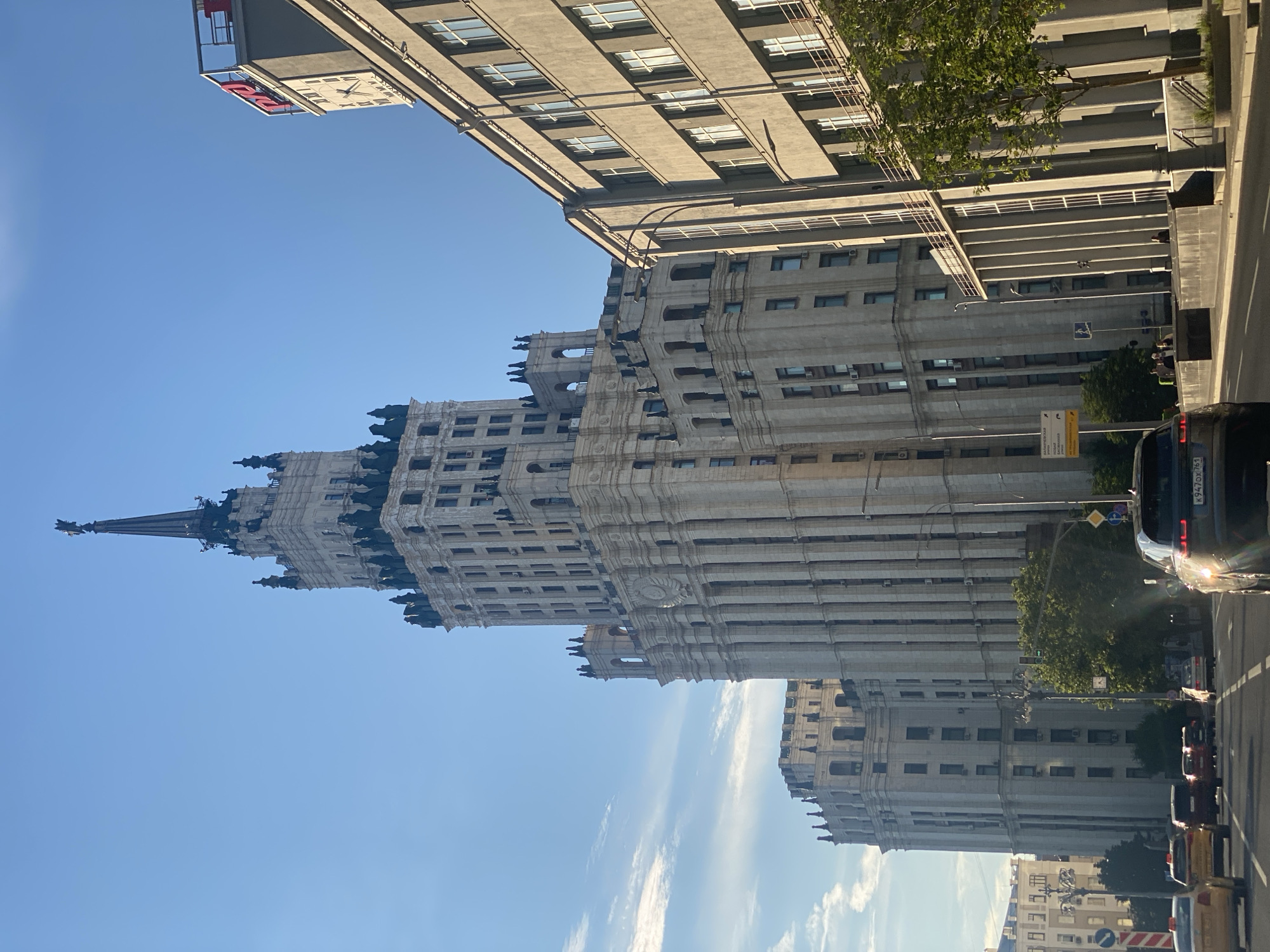 An external, eye-level, medium shot captures a sunlit street scene in Moscow, Russia, focusing on the monumental Stalinist Empire style architecture of the Ministry of Foreign Affairs building, one of the city's famous Seven Sisters skyscrapers. The building, with its distinctive spire and decorative elements, dominates the frame. Adjacent to it is another similar, though shorter, building. A modern gray office building stands to the right, featuring a prominent clock face and a red illuminated sign with Cyrillic lettering, possibly "РЖД" (Russian Railways). The sky is a clear, bright blue with a few wispy clouds. A multi-lane road in the foreground has several cars, including a yellow taxi and a dark SUV, driving or waiting. A few pedestrians are visible on the sidewalk. Green trees are interspersed throughout the scene, adding a touch of nature to the urban environment. The lighting suggests it is daytime, likely morning or afternoon, with sunlight casting shadows and highlighting the architectural details.