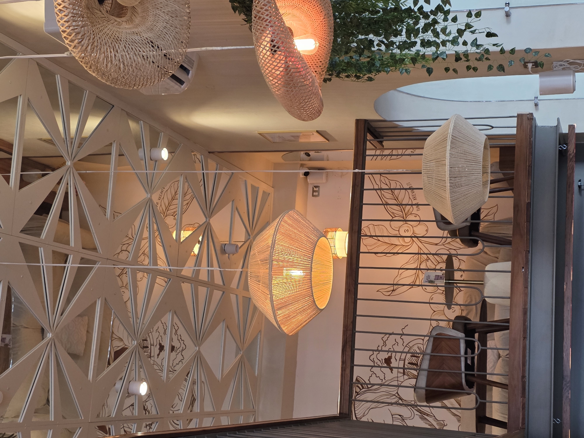 The image captures an interior scene, possibly a cafe or restaurant, in Tijuana, Mexico. The camera angle is tilted, presenting an unconventional perspective.
The scene is illuminated by several hanging light fixtures, crafted from materials like wicker and rope, and emitting a warm, orange glow. Some of the lamps are round and flat, while others are elongated and hexagonal.
A geometric, decorative screen with triangular cutouts is visible on one side, while the opposite side features a metal railing and a wall mural depicting coffee beans and other design elements. Seating is visible behind the railing, suggesting a dining area.
Green foliage is draped across the ceiling in the upper part of the frame, adding a touch of nature to the scene. Other ceiling-mounted fixtures and structural elements can be seen. The overall ambiance is warm and inviting.