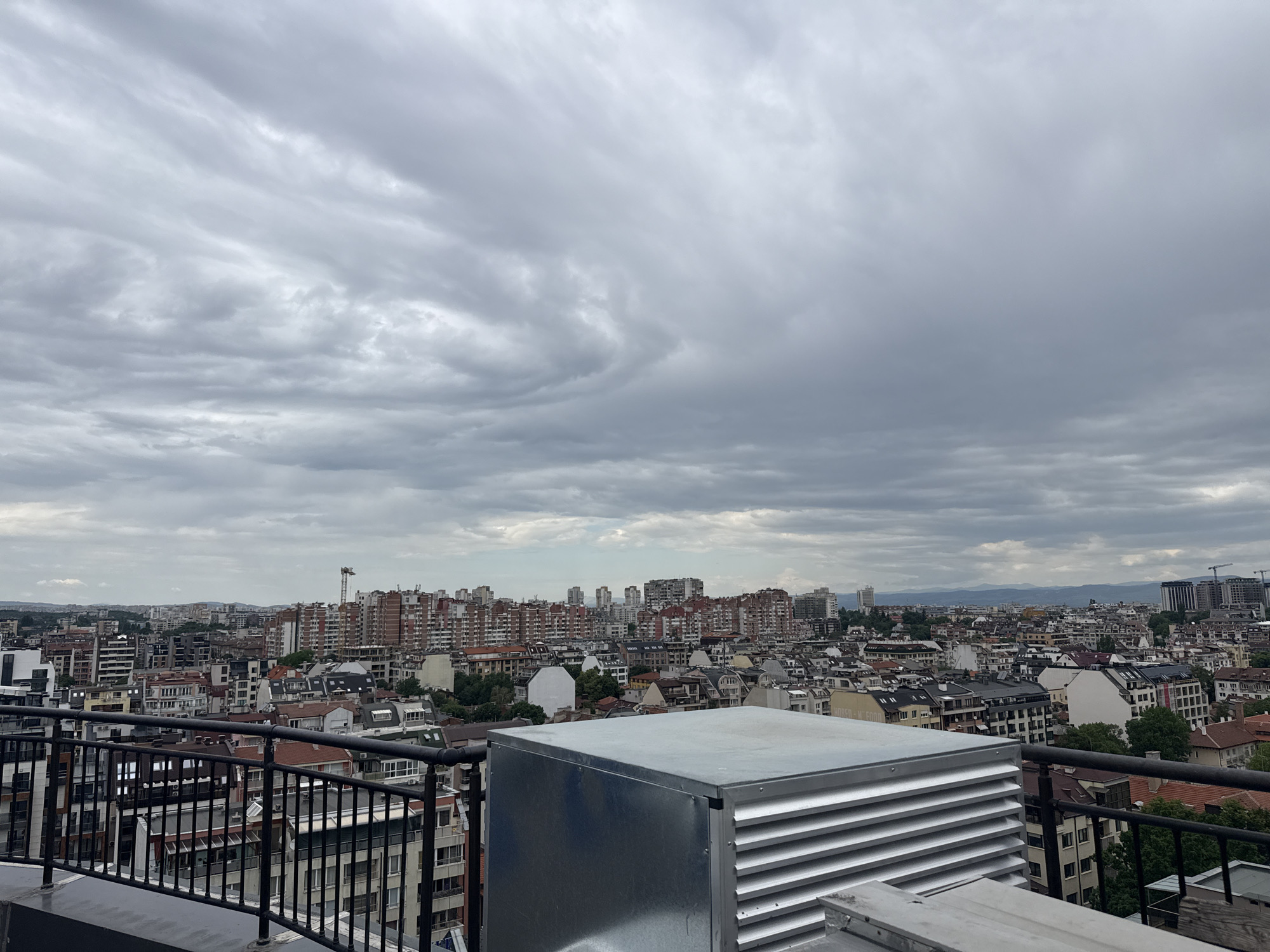 The image shows a view of Sofia, Bulgaria from a high vantage point, likely a rooftop. The foreground includes a metal structure with ventilation slats and a metal railing. The cityscape stretches out behind, with a mix of residential and commercial buildings, many with red-tiled roofs. A construction crane is visible in the distance. The sky is overcast with swirling gray clouds. No people are visible in the scene. The overall impression is one of a densely populated urban environment under a gloomy sky.