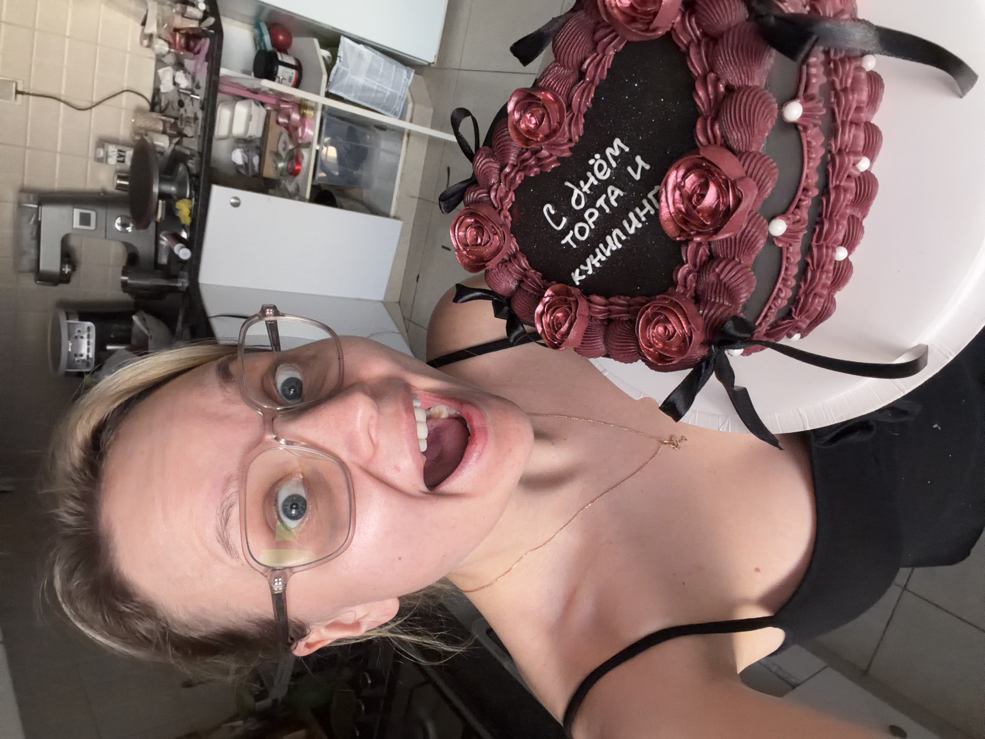 A woman wearing glasses and a black tank top is holding a cake. The cake is decorated with maroon frosting roses, black ribbons, and white pearls. There is writing in Russian on the cake. The location is a kitchen with white cabinets and appliances.