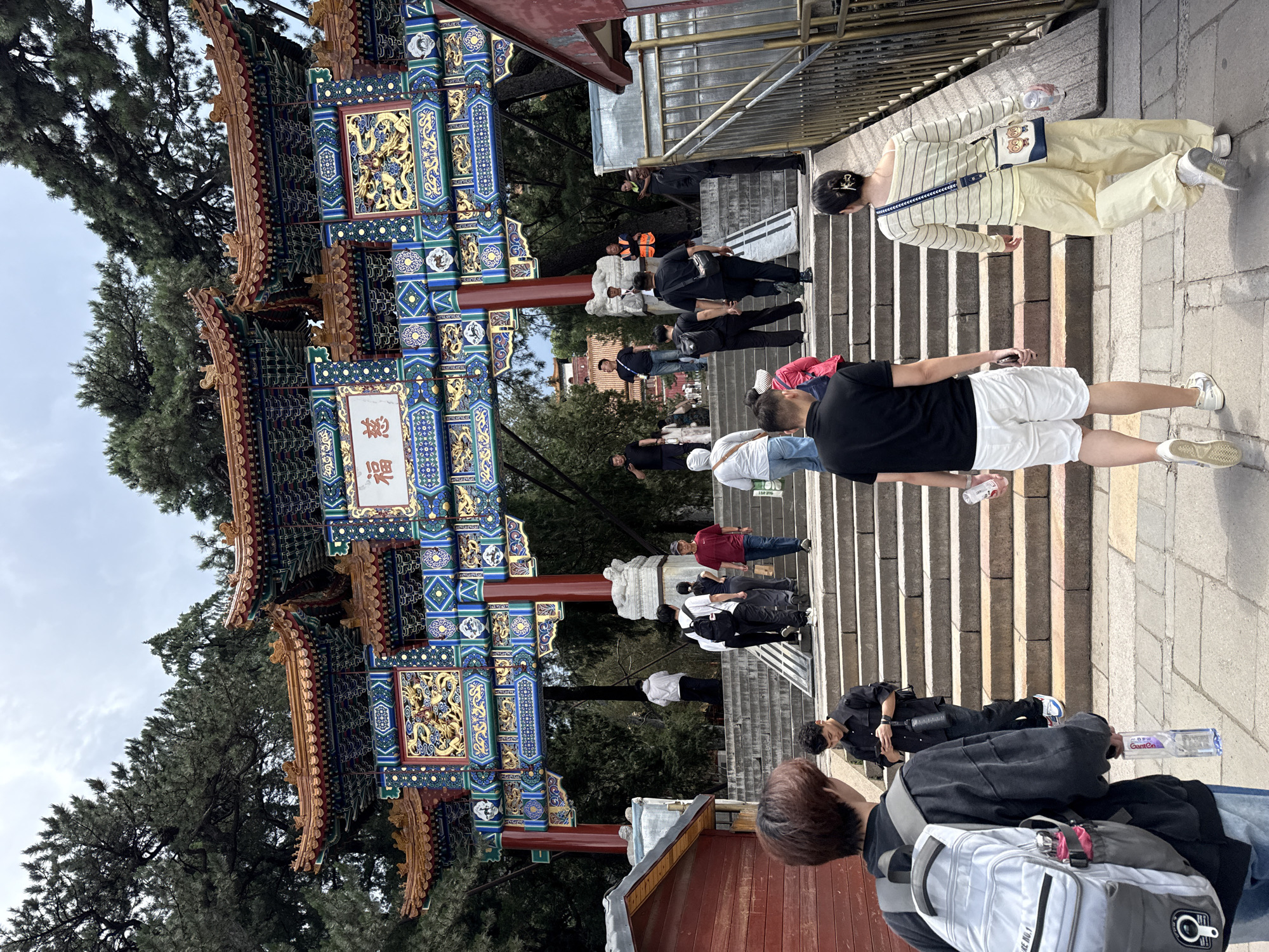 This image captures the entrance to the Fragrant Hills Park (Xiangshan Gongyuan) in Haidian District, Beijing, China, specifically depicting the Cífúmén (Gate of Benevolent Fortune) archway.
The prominent central feature is a large, ornate traditional Chinese archway, commonly known as a paifang or pailou. It is elaborately decorated with multiple tiered roofs featuring upturned eaves and intricate patterns in red, blue, green, and gold. Gold relief carvings, possibly depicting dragons or mythical creatures, are visible on the friezes and panels. A central white plaque on the archway bears two red Chinese characters reading "慈福" (Cífú), meaning "Benevolent Fortune" or "Compassionate Blessing." The archway is supported by large red columns.
In the foreground, a wide, multi-tiered stone staircase ascends towards and through the archway. Numerous individuals, estimated between 20 and 30, are present on the stairs, mostly walking upwards, suggesting entry into the park. People are casually dressed, with a mix of light and dark clothing.
- In the immediate foreground on the left, a person with short brown hair, wearing a dark top and light blue jeans, carries a light gray backpack with a pink water bottle and holds a clear plastic water bottle.
- To their right, a male in a dark long-sleeved shirt and dark pants is looking at a smartphone.
- On the far right foreground, a person in a striped long-sleeved top and loose light yellow pants carries a small white and blue shoulder bag with an owl design and a clear cup. Another male in a black polo shirt and white shorts walks beside them.
Further up the stairs, a diverse group of individuals includes a person in an orange safety vest on the right side of the archway, suggesting staff or security. Other individuals appear to be visitors.
The environment is characterized by dense, dark green evergreen trees (likely pines) visible behind and surrounding the archway, indicating a park or mountainous setting. The sky is overcast with a uniform light gray cloud cover, resulting in diffuse lighting. Small, single-story structures painted red are positioned on both sides of the staircase in the foreground, possibly functioning as ticket booths or vendor stalls. A glimpse of another traditional Chinese-style building with a red roof is visible through the archway in the distance.
